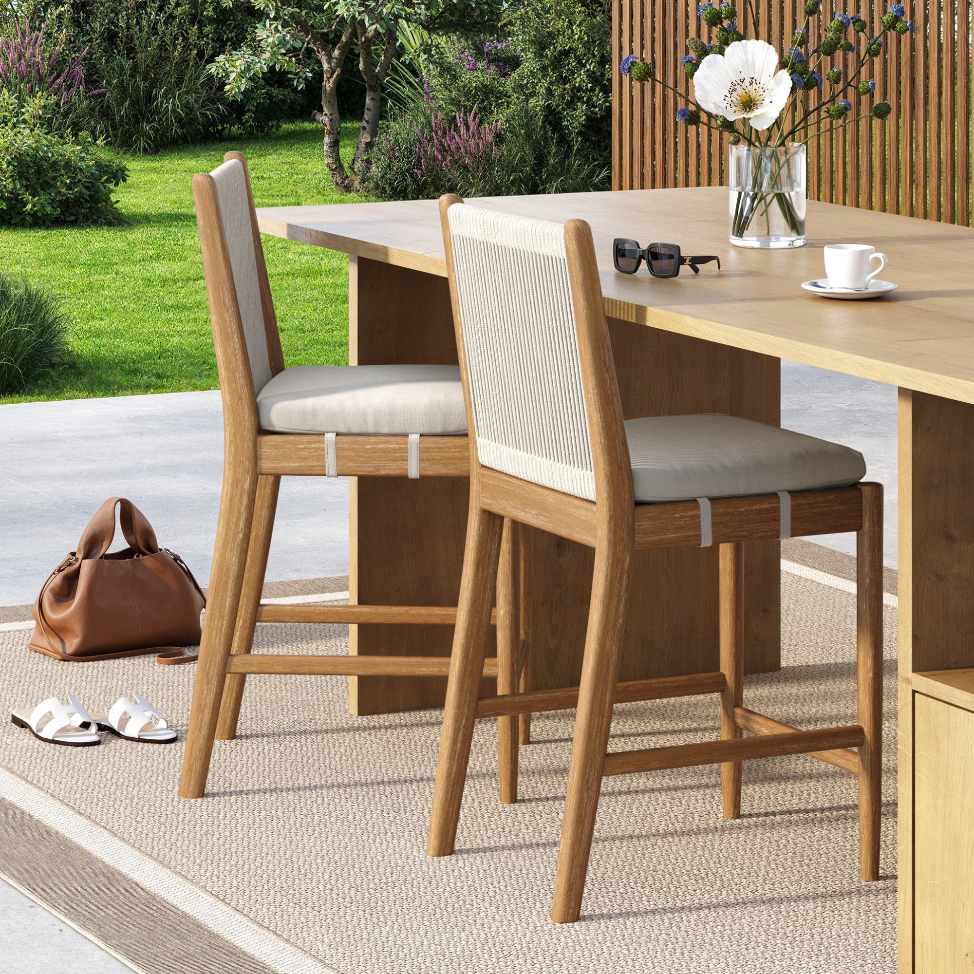 Two Nathan James Outdoor Wood & Rope Counter Height Bar Stools in brushed light brown stand by a light wood table outdoors, decorated with a vase of flowers, cup, sunglasses, and book. A brown bag and white sandals rest on a beige rug nearby.