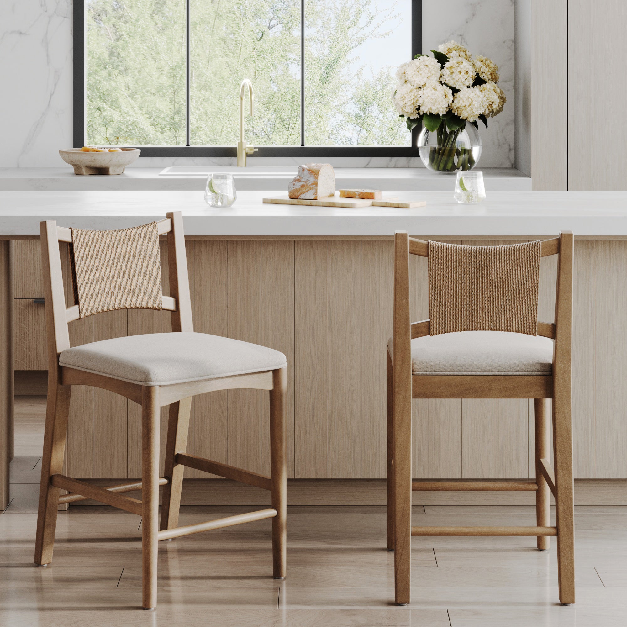 A set of two Nathan James Seagrass Back Arched Footrest Counter Height Bar Stools sits in a modern kitchen with light wood cabinets, a white countertop, white flowers in a vase, and a window overlooking lush greenery.