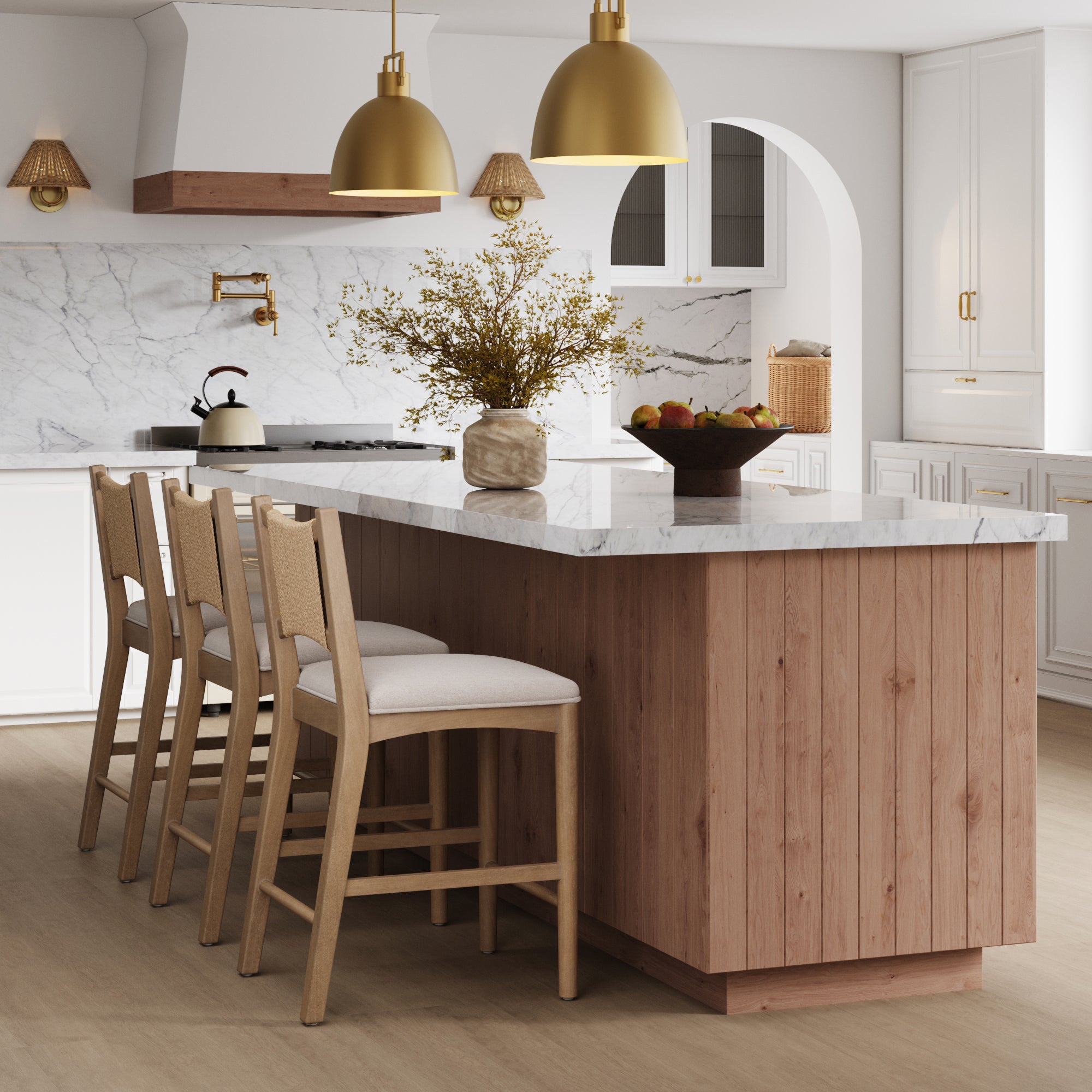 A modern kitchen features a wooden island, marble countertops, gold pendant lights, white cabinets, a vase with branches, a fruit bowl, and three Nathan James Seagrass Back Arched Footrest Counter Height Bar Stools in a bright neutral palette.