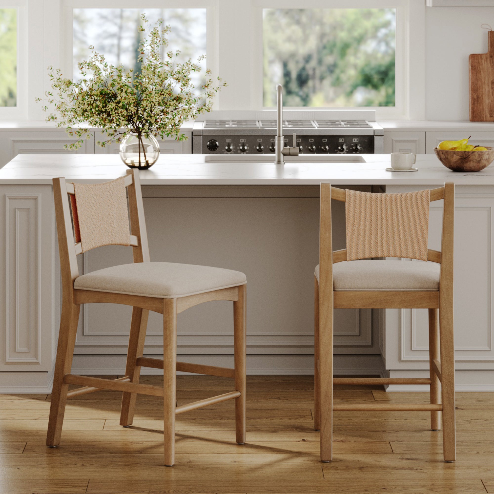 Two Nathan James Seagrass Back Arched Footrest Counter Height Bar Stools with woven backrests and light fabric seats stand at a white kitchen island, topped with a sink, flowers, bananas, and a cup beneath large windows.