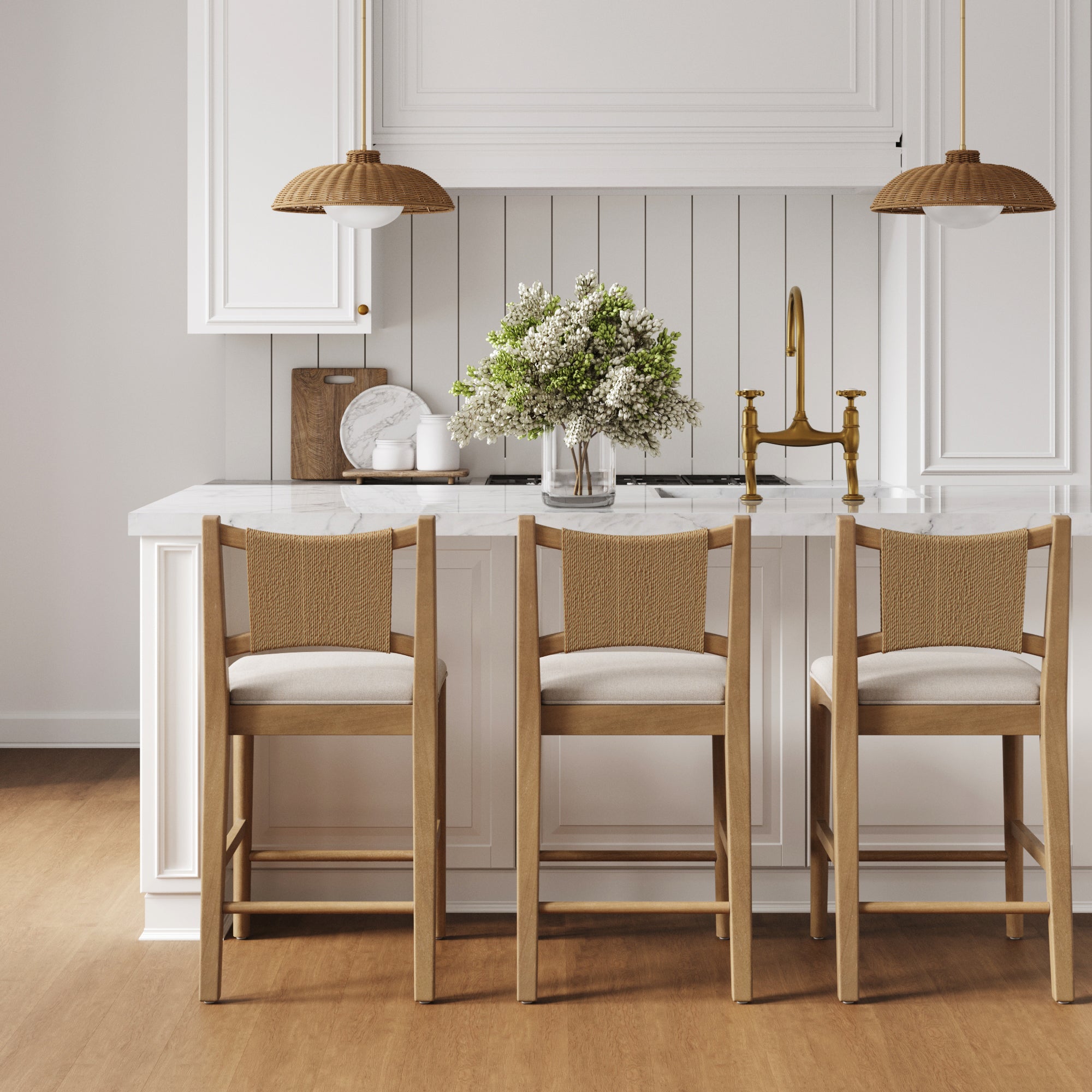 A modern kitchen with a white island, three Nathan James Seagrass Back Arched Footrest Counter Height Bar Stools, brass fixtures, white cabinets, hanging rattan lights, and a vase of greenery on the countertop.