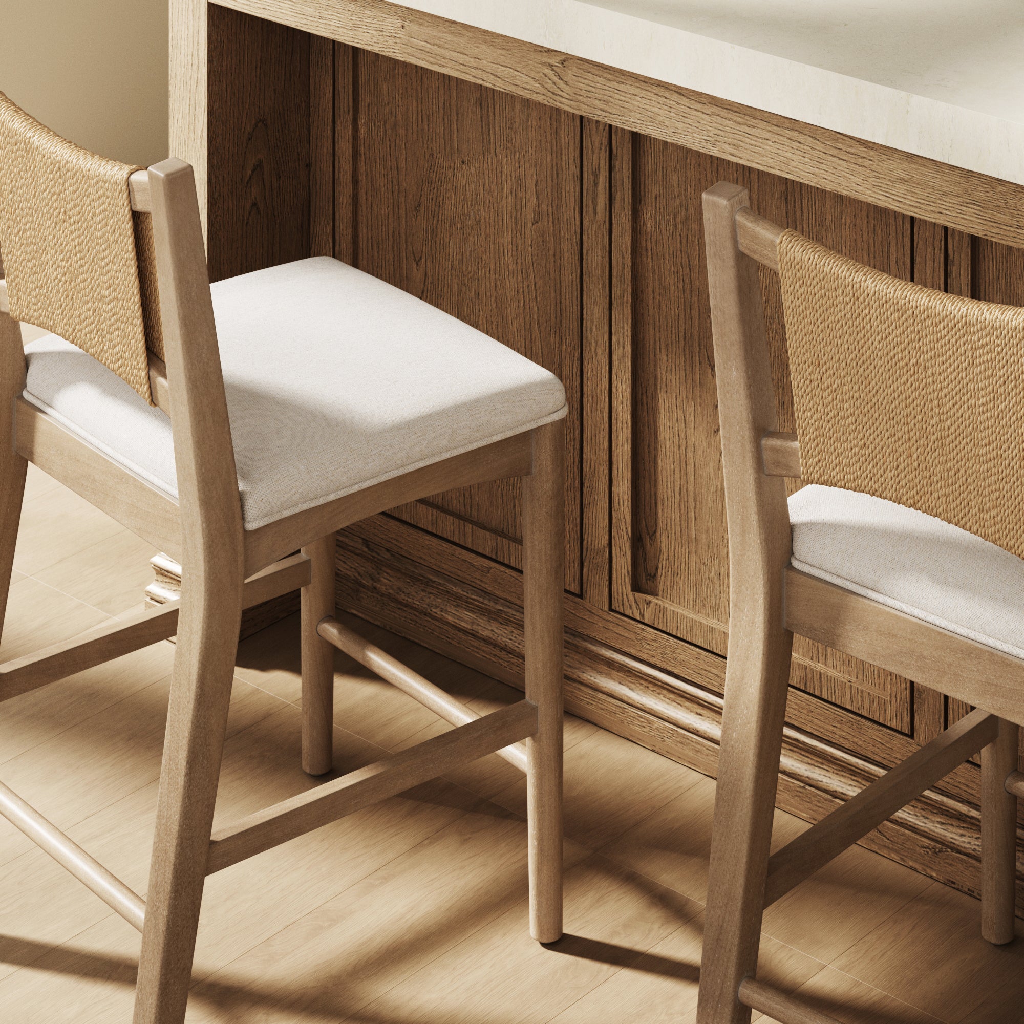 Two wooden bar stools with light-colored padded seats and woven backrests are placed at a wooden kitchen counter with a light countertop. The scene has a warm, natural, and minimalist aesthetic.