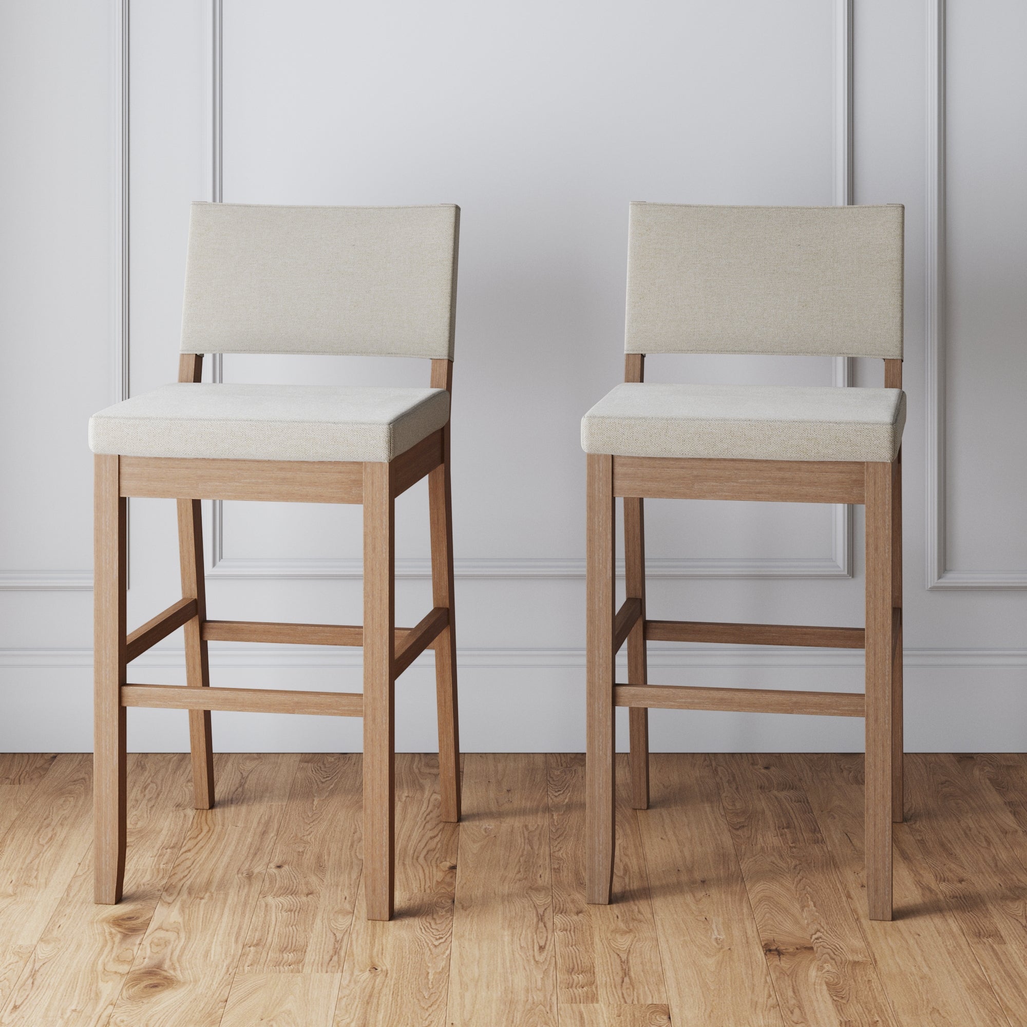 A set of two Nathan James Wood Upholstered Bar Stools in light brown adds a modern farmhouse touch, placed side by side on a wooden floor against a white paneled wall.