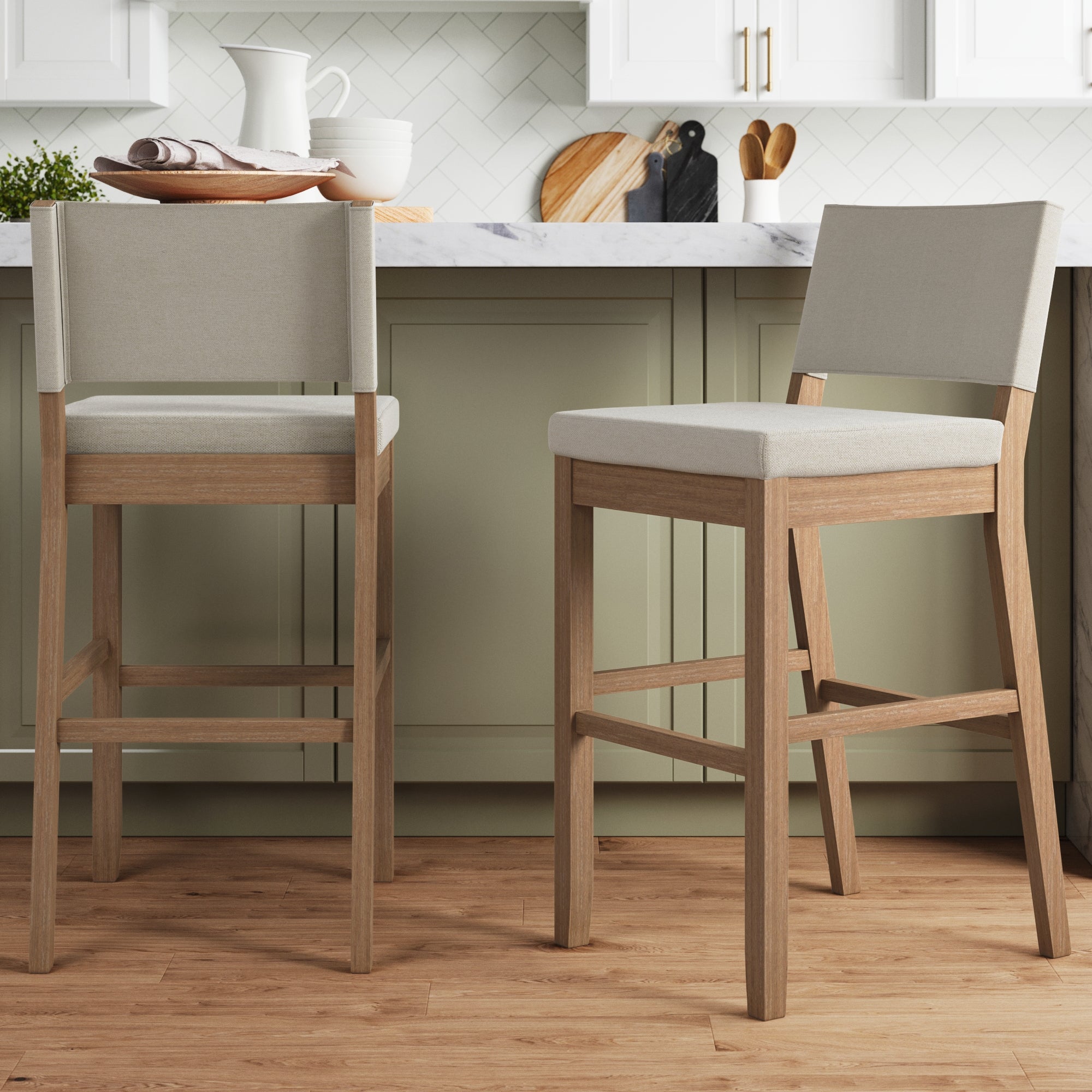 Two Nathan James Wood Upholstered Bar Stools in light brown (set of 2) are placed in a modern kitchen with green cabinets, marble countertop, and white backsplash; kitchen items and cutting boards rest on the counter.