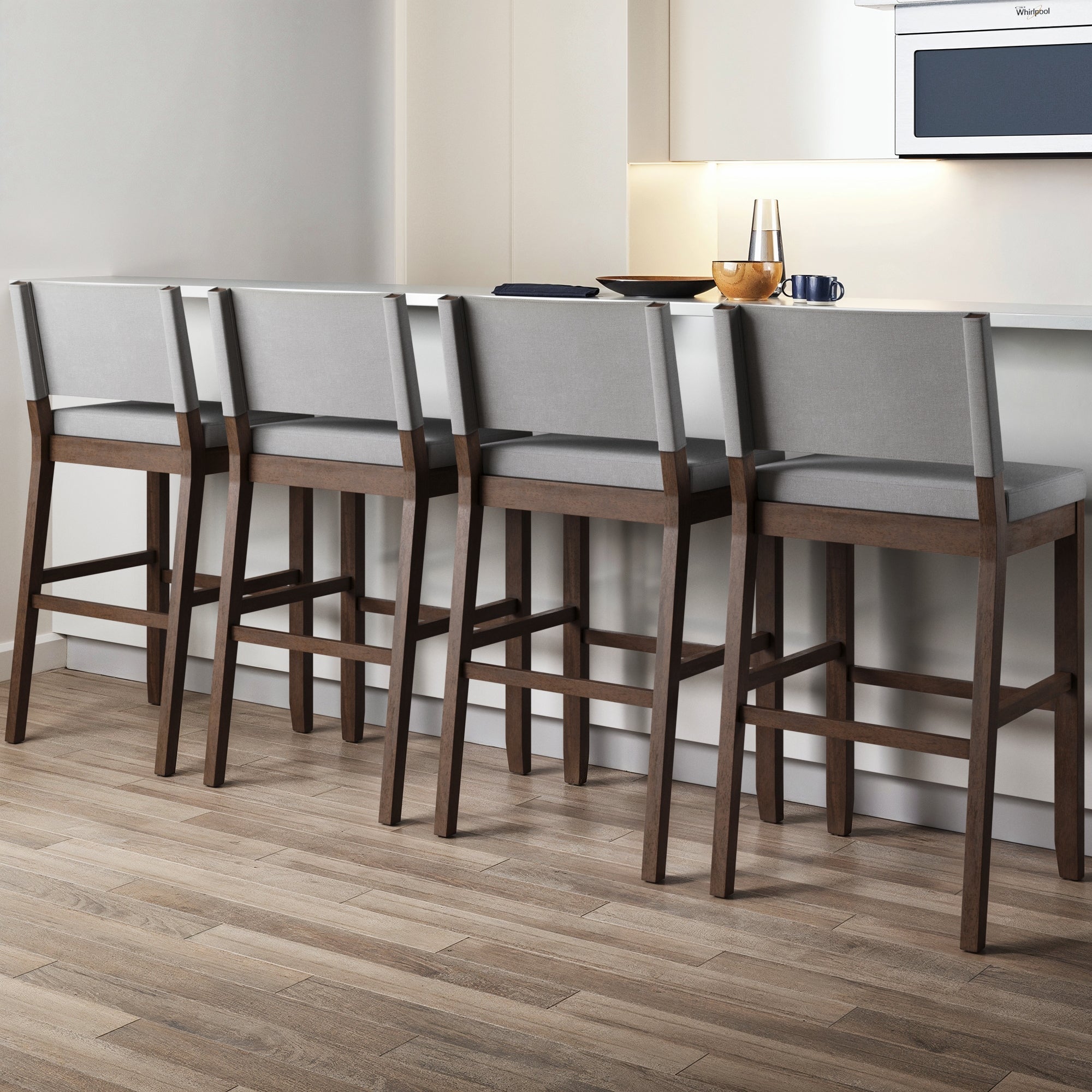 Four Nathan James Wood Upholstered Sling Back Bar Stools in gray are lined up at a white kitchen counter with light wood flooring. The set of 4 features solid wood frames and upholstered seats; the counter holds a pitcher, glass, bowl, and plate.