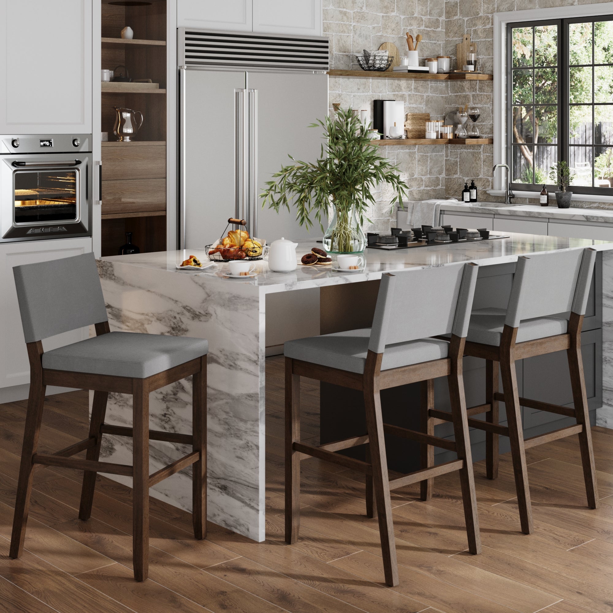 Modern kitchen with a marble island, three Nathan James Wood Upholstered Sling Back Bar Stools in gray, wooden floor, built-in oven and refrigerator. A plant and breakfast items sit on the island; large window provides natural light and views.