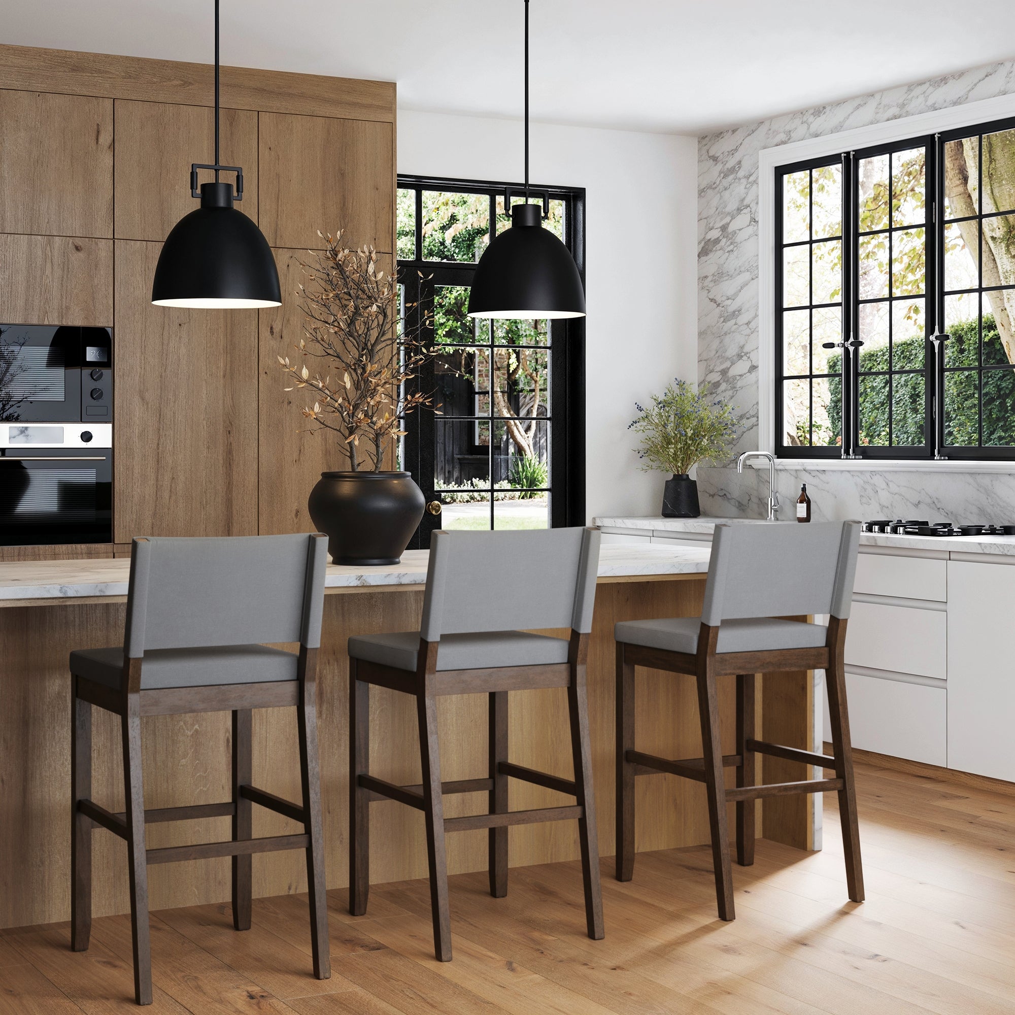 Modern farmhouse kitchen with solid wood cabinets, marble countertops, and three Nathan James Wood Upholstered Sling Back Bar Stools in dark brown gray. Black pendant lights hang above the island with a black vase. Large windows fill the space with natural light.
