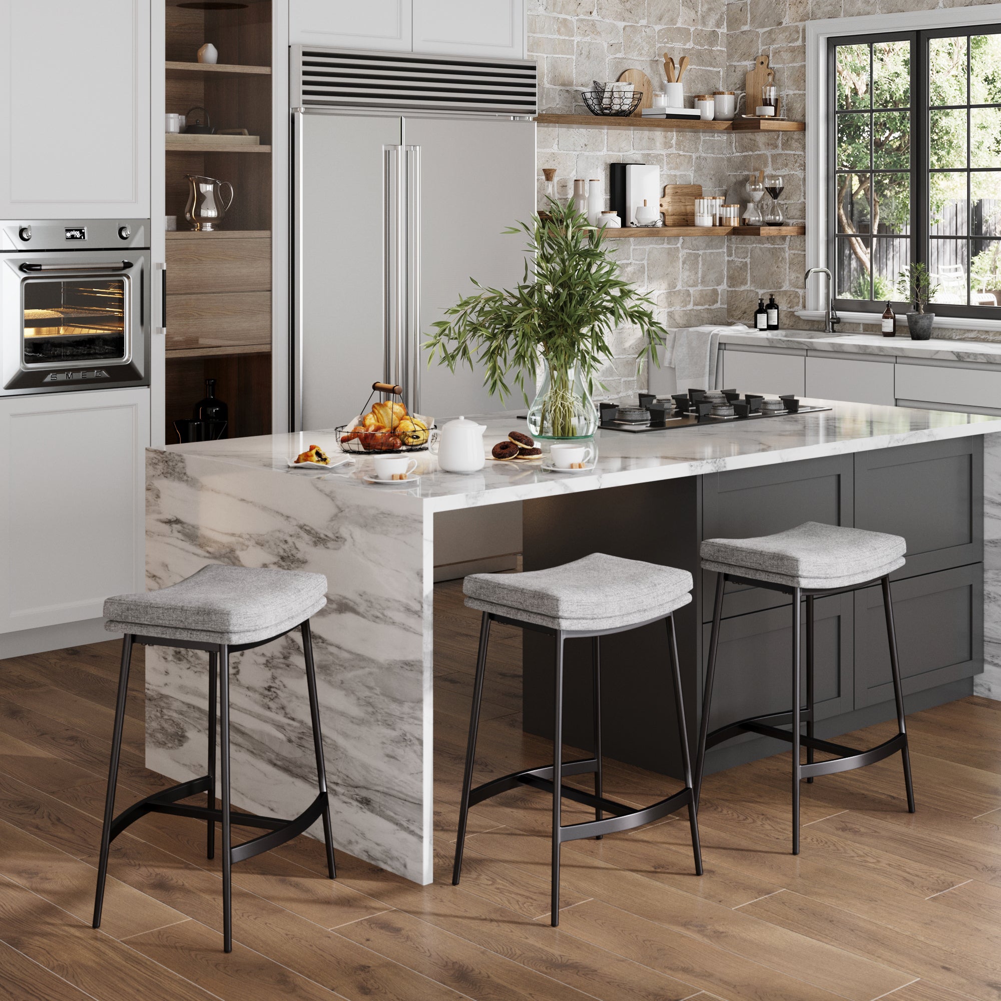 Modern farmhouse kitchen with a marble island, three Nathan James Upholstered Counter Height Saddle Bar Stools in Gray, built-in oven, stainless refrigerator, wood floors, open shelves with dishes, and a vase of greenery on the countertop by a sunny window.