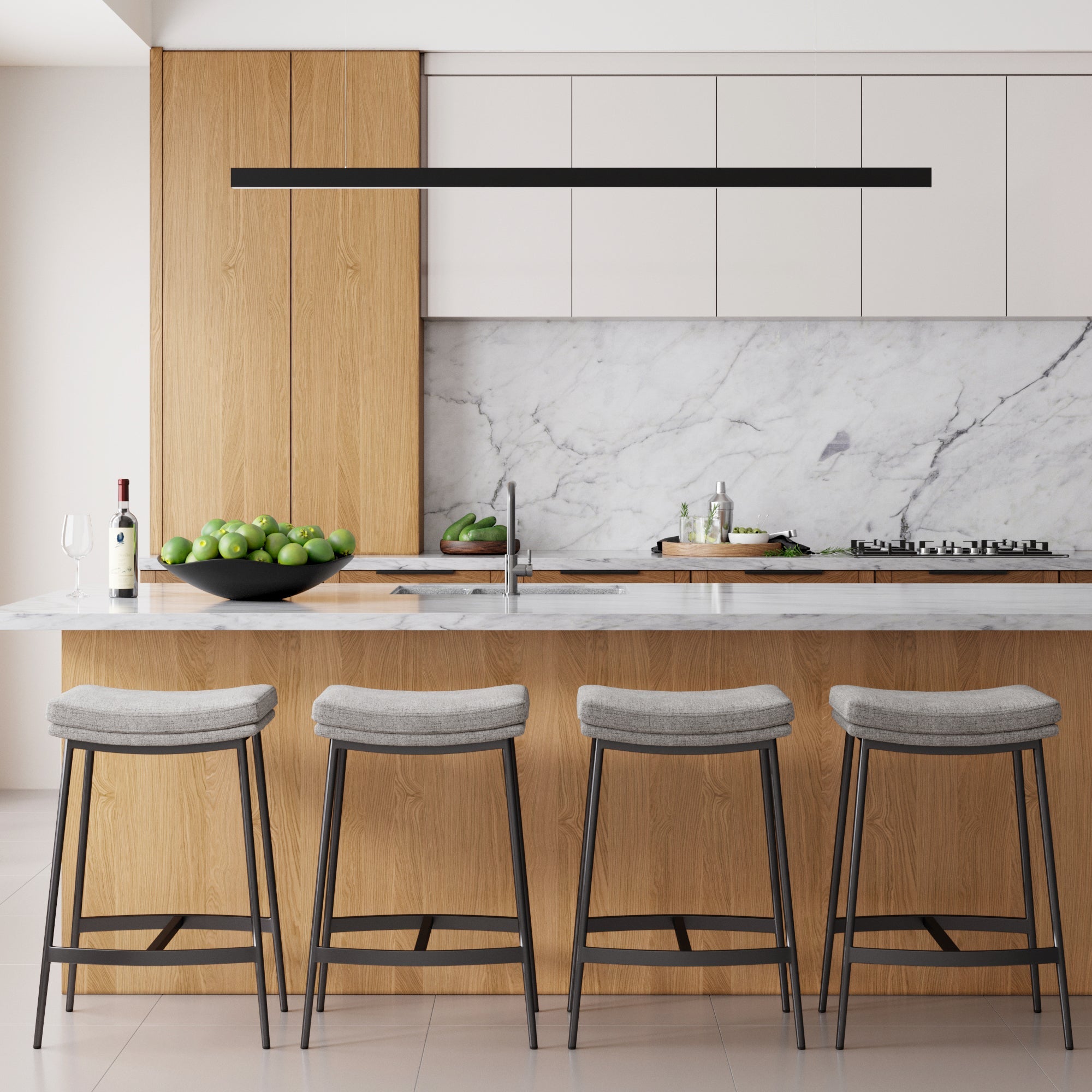 A modern farmhouse kitchen features wood cabinets, a marble backsplash and countertop, four Nathan James Upholstered Counter Height Saddle Bar Stools in gray, a bowl of green apples, a wine bottle with glass, and minimalist decor under a linear black light fixture.