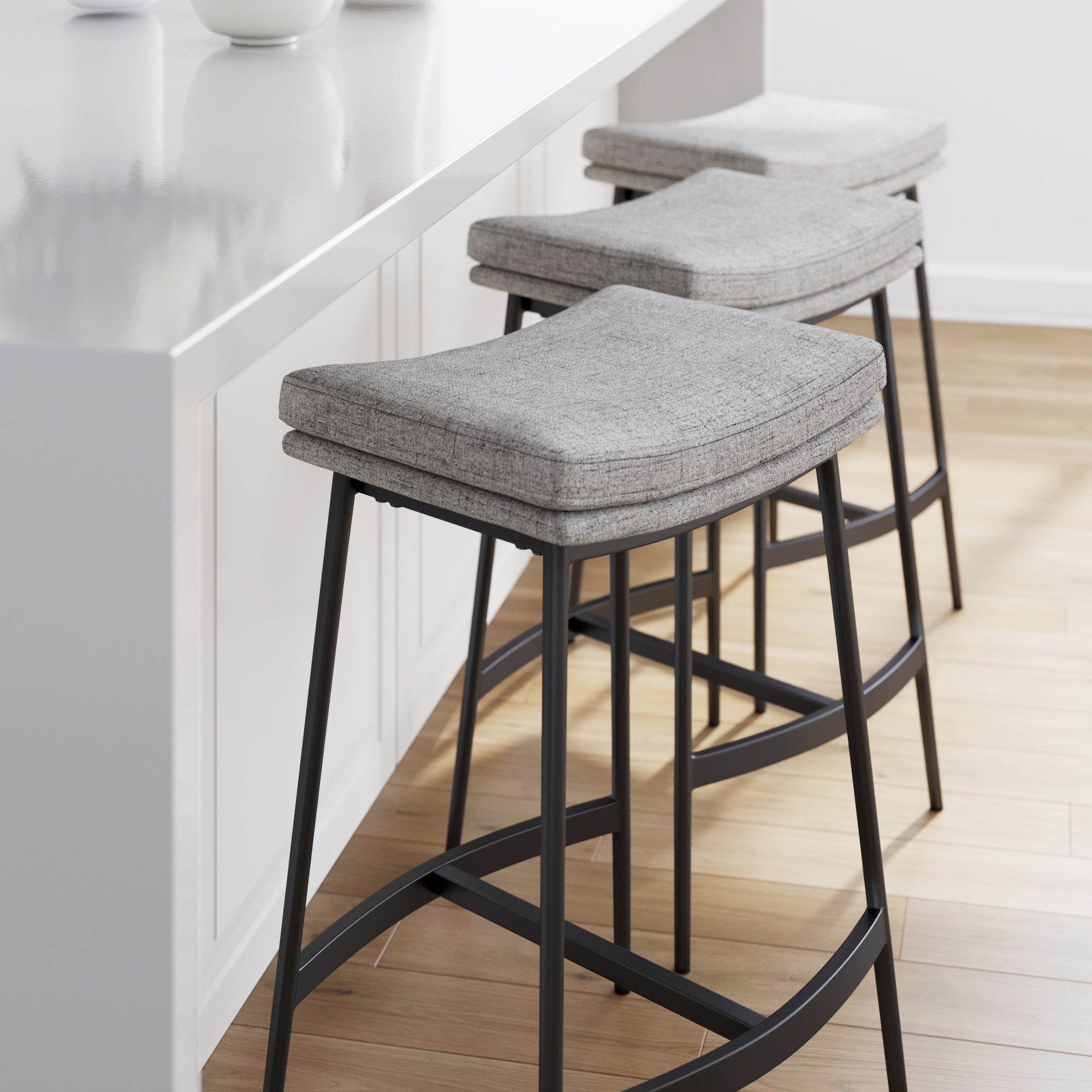 Three modern bar stools with gray upholstered seats and black metal legs are lined up beside a white kitchen island on a light wood floor in a bright, minimalist kitchen.
