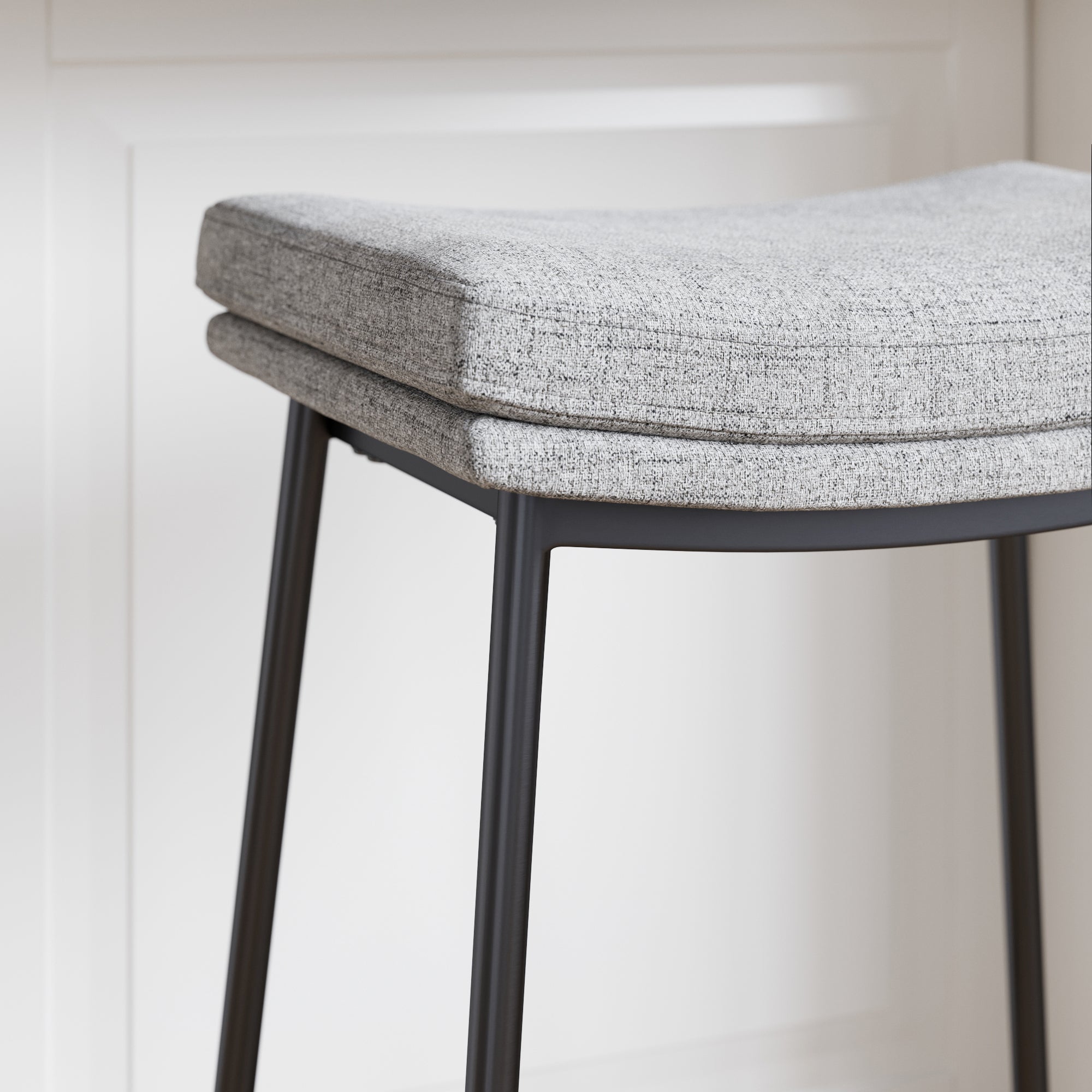 A close-up of a modern stool with a soft, gray upholstered cushion and slender black metal legs, positioned in front of a white paneled background.