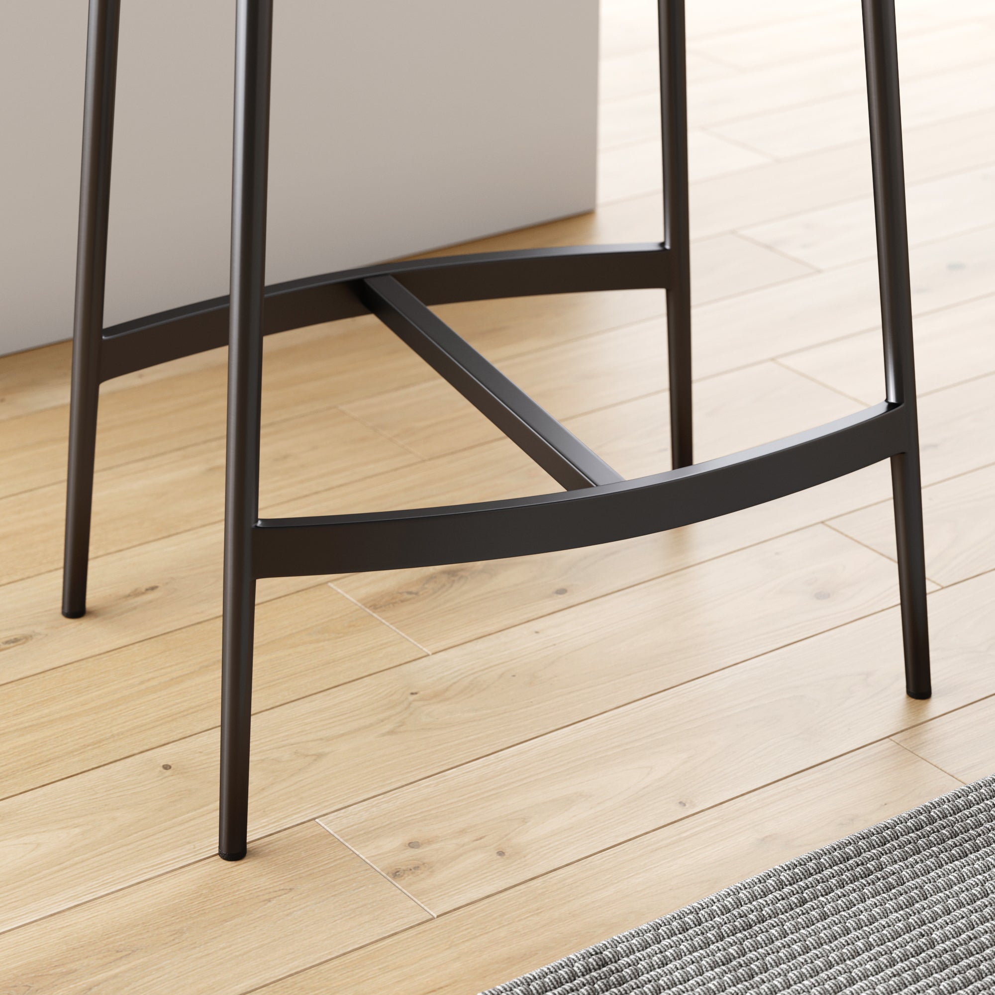 Close-up of the black metal legs and footrest of a bar stool on light wood flooring, next to a textured gray rug and a white wall or counter.