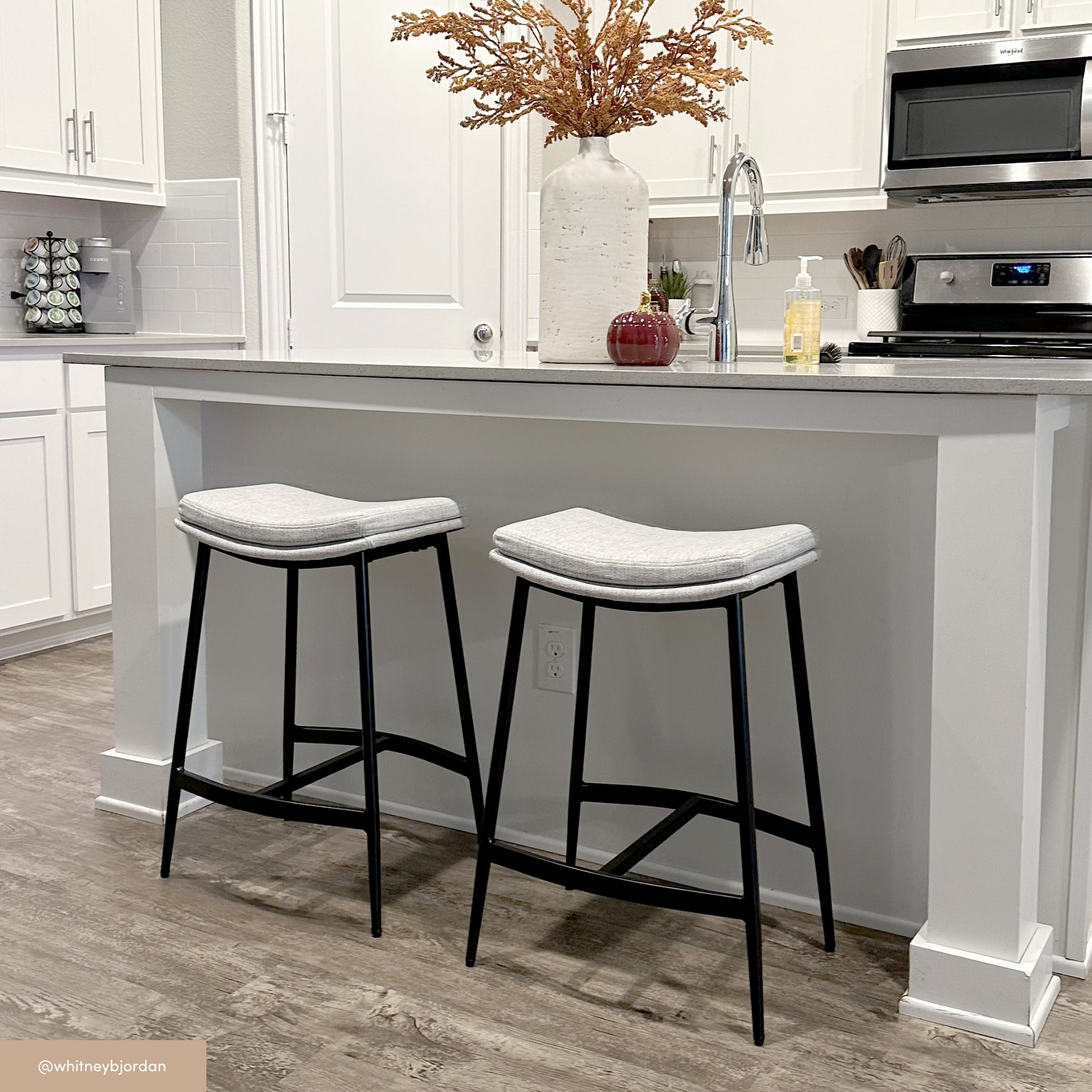 A modern kitchen with white cabinets and a light gray island, featuring two gray cushioned bar stools with black legs. A large vase with dried branches sits on the counter, along with a small red decorative item and soap.
