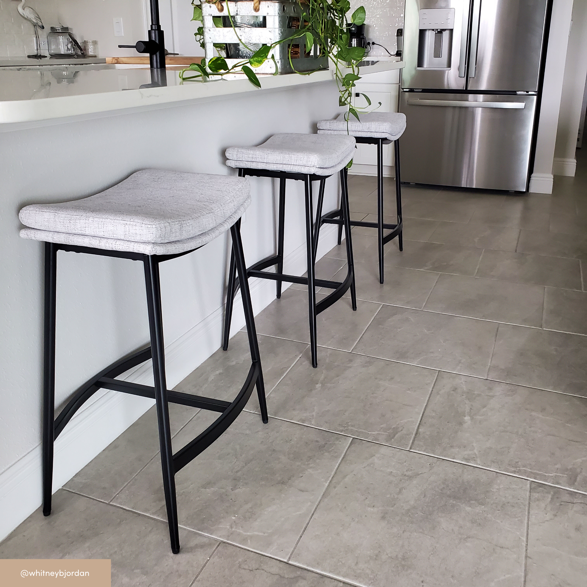 Three modern gray bar stools with black metal legs are lined up at a white kitchen counter. The floor is tiled, and a stainless steel refrigerator is visible in the background. A plant hangs over the counter.