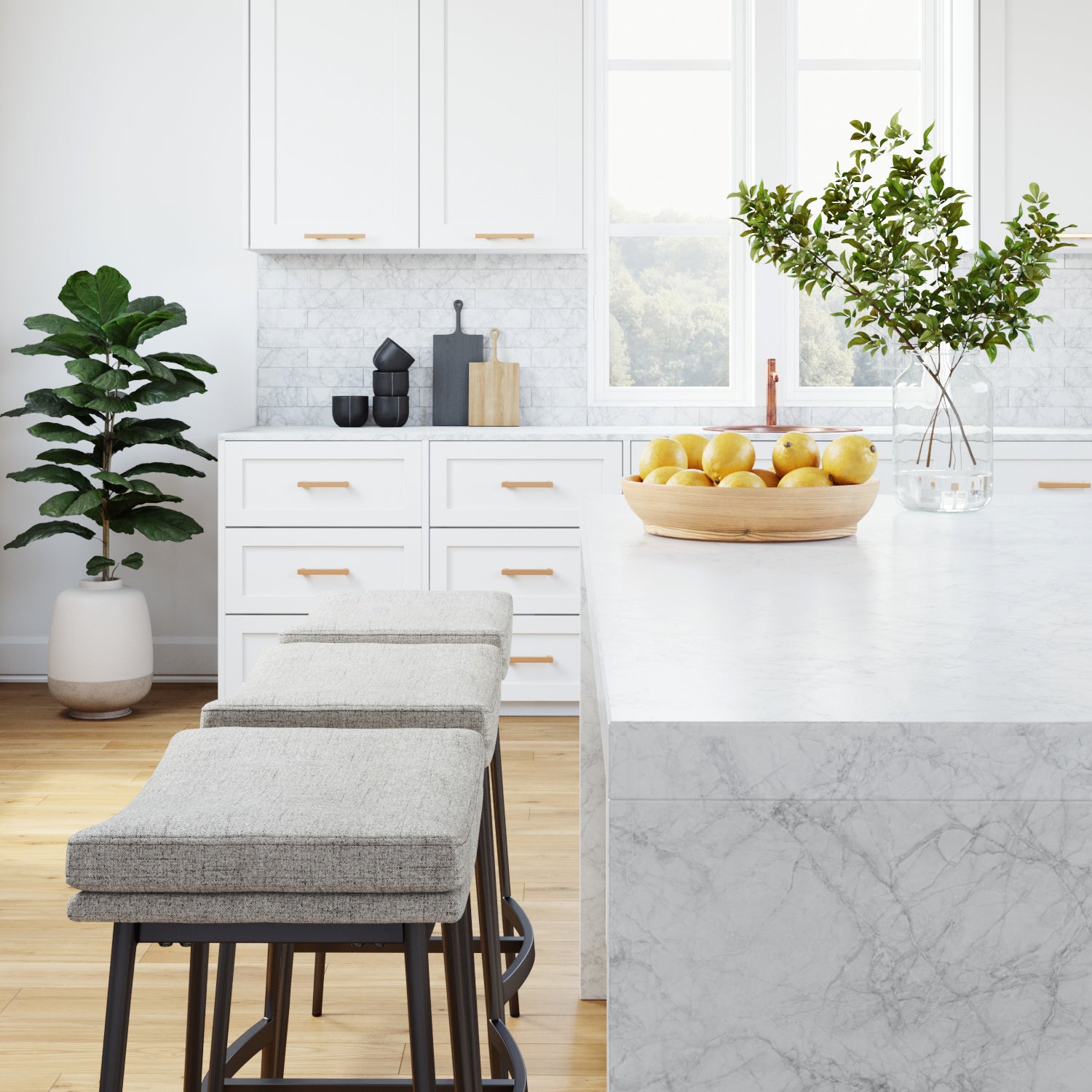 A modern kitchen with white cabinets, marble countertops, two cushioned stools, a wooden bowl of apples on the island, a tall green plant, and large windows letting in natural light.