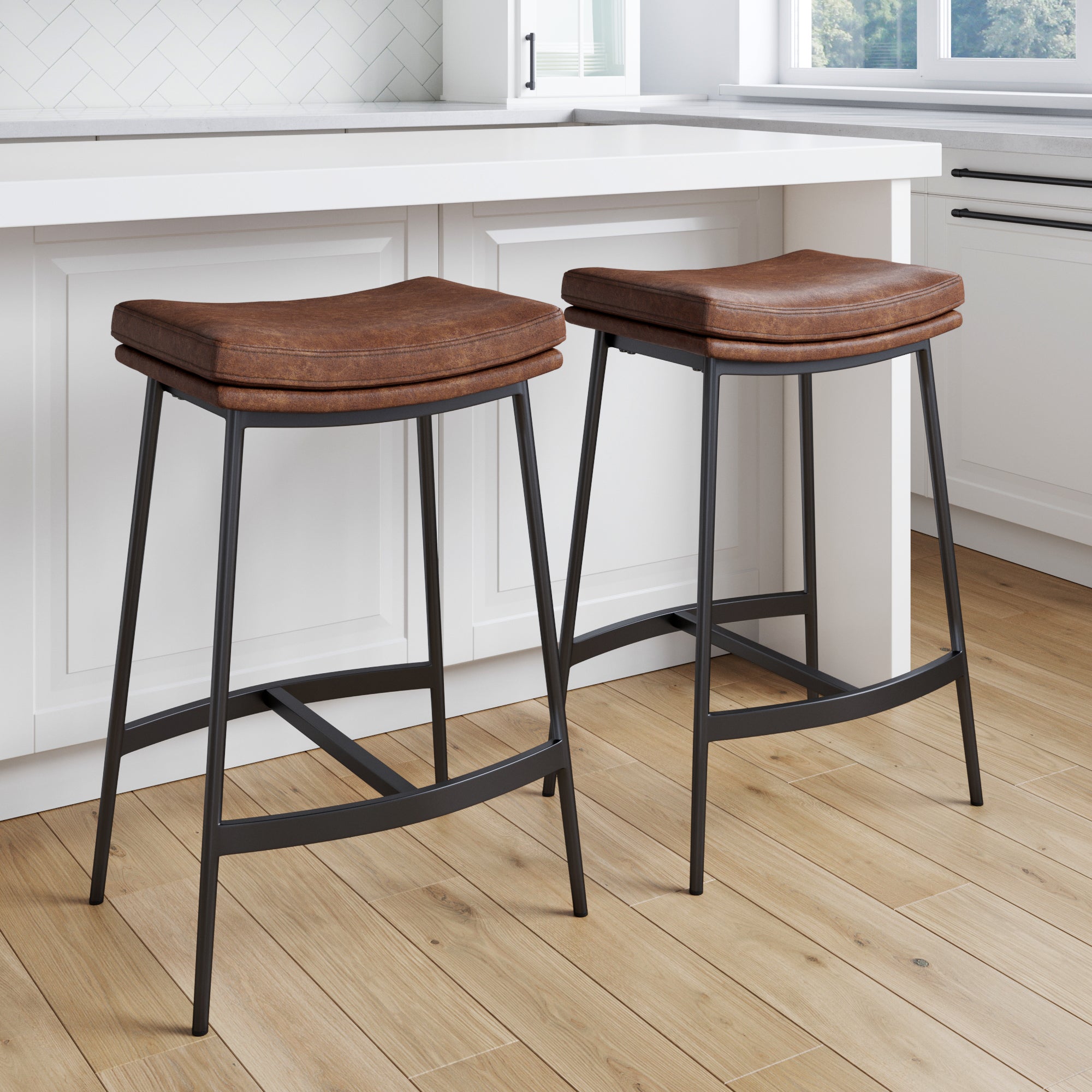 Two brown leather bar stools with black metal legs are placed in front of a white kitchen counter on a light wood floor. The kitchen has white cabinets and natural light coming through a window.