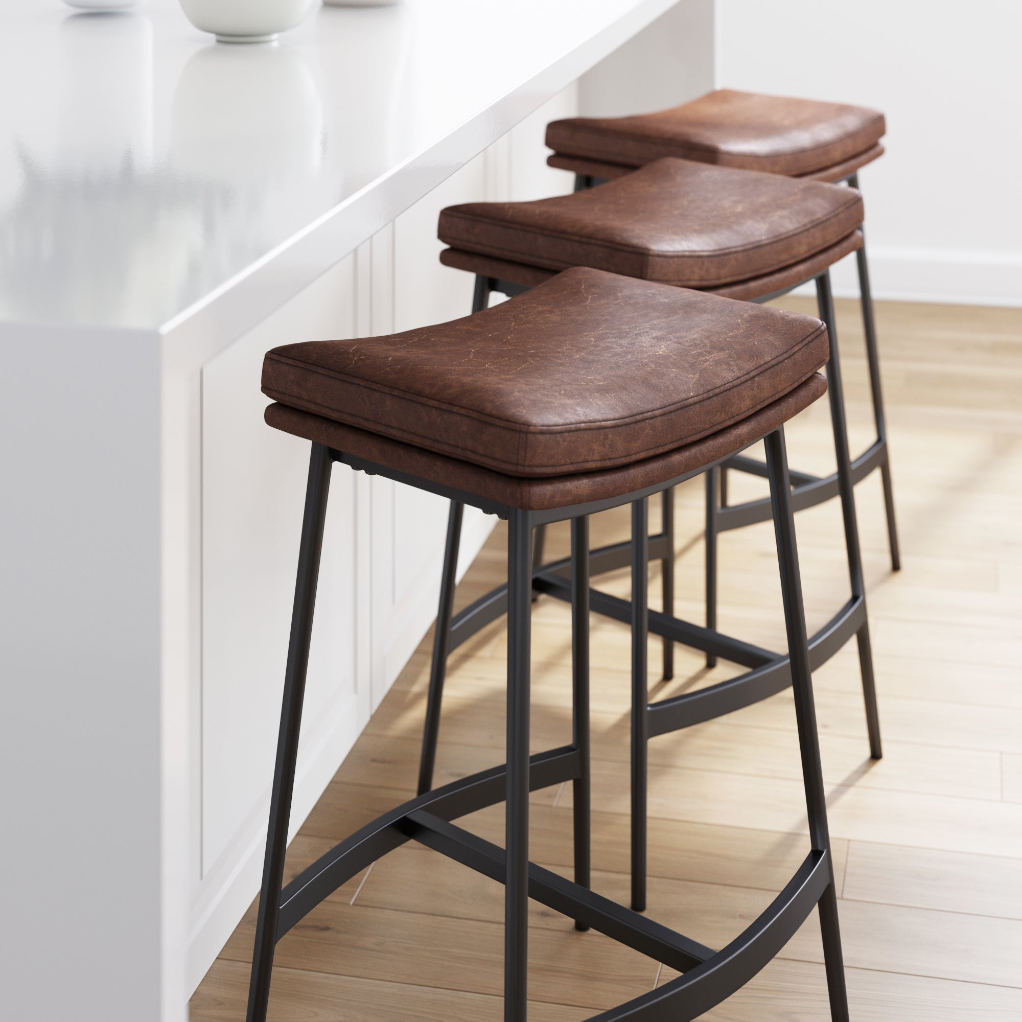 Three brown leather bar stools with black metal legs are lined up beside a white kitchen island on a light wood floor in a modern, minimalistic kitchen.