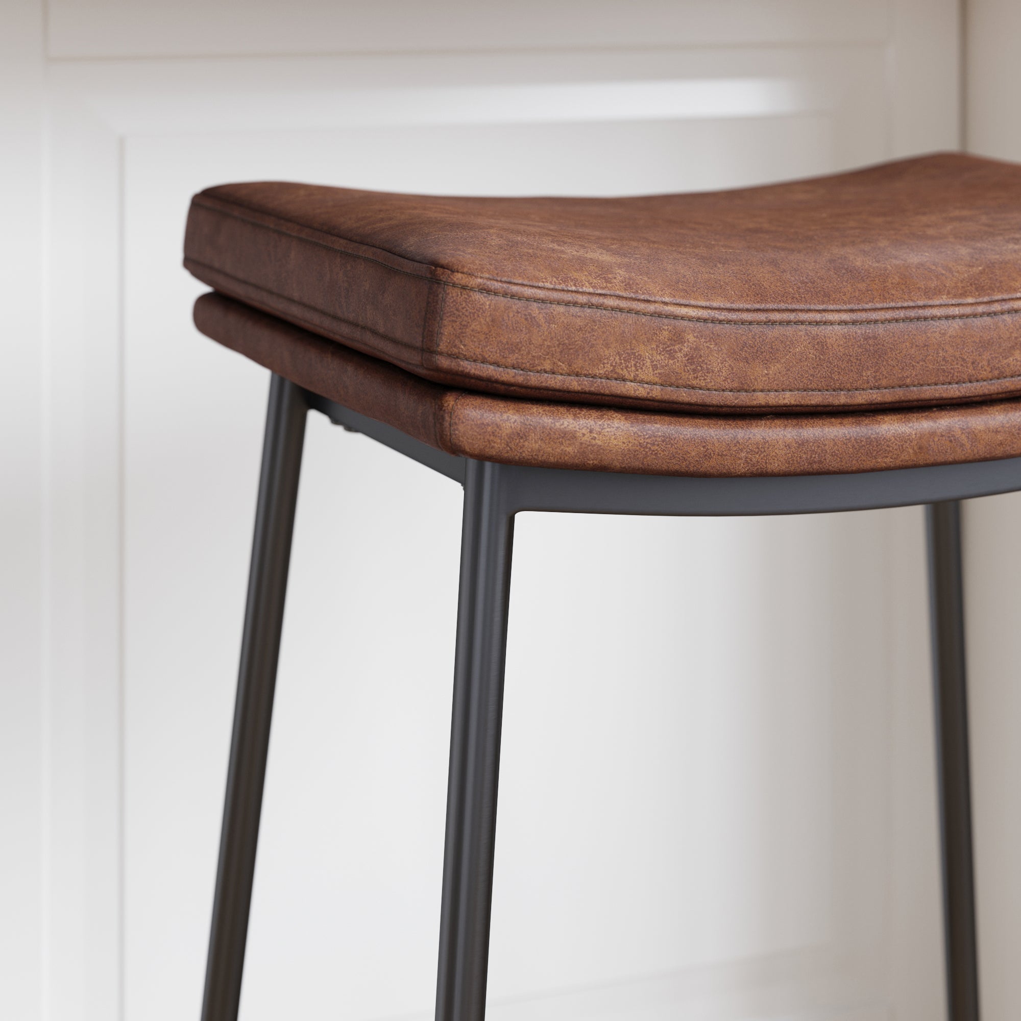 A close-up of a modern barstool with a brown cushioned leather seat and slim black metal legs, positioned in front of a white panel wall.