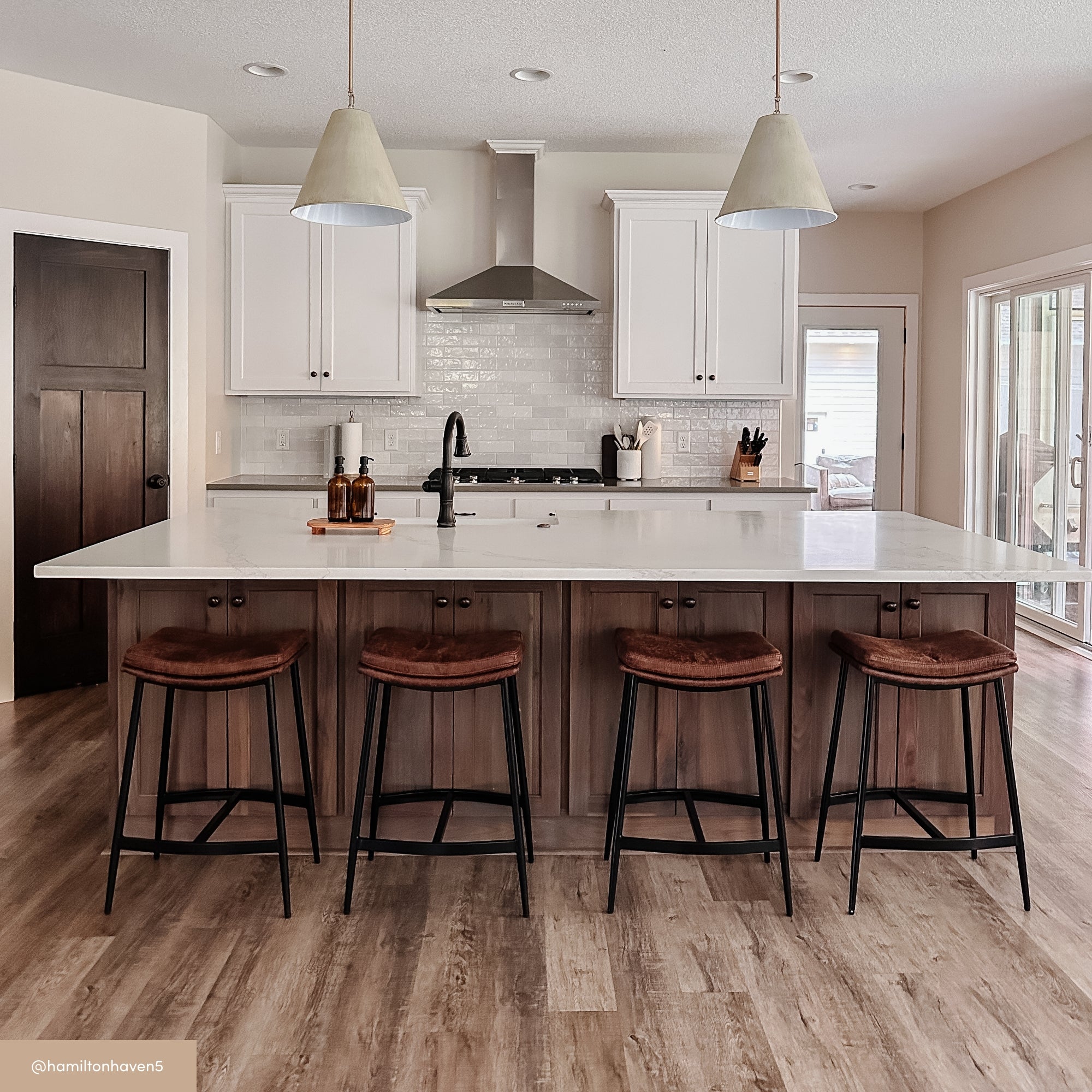 A modern kitchen with a large island, four brown cushioned stools, white cabinets, a stainless steel range hood, and pendant lights. The space has wood flooring and sliding glass doors letting in natural light.
