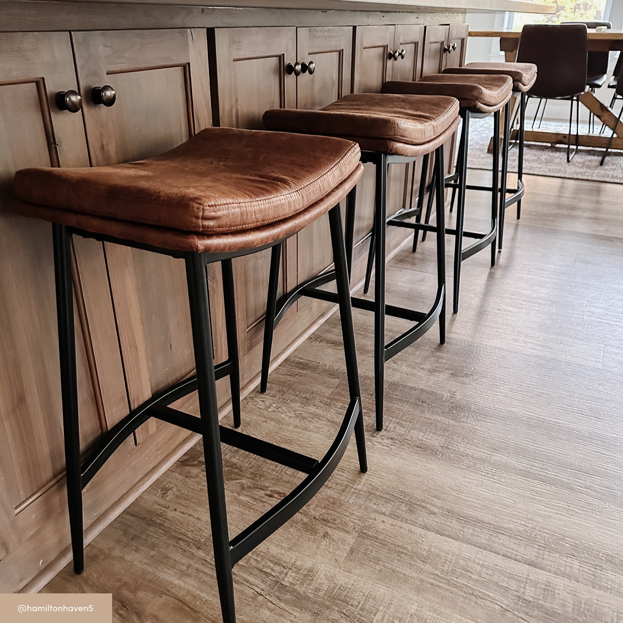 Three brown leather bar stools with black metal legs are lined up at a wooden kitchen island on a light wood floor, with sunlight streaming in from a nearby window.