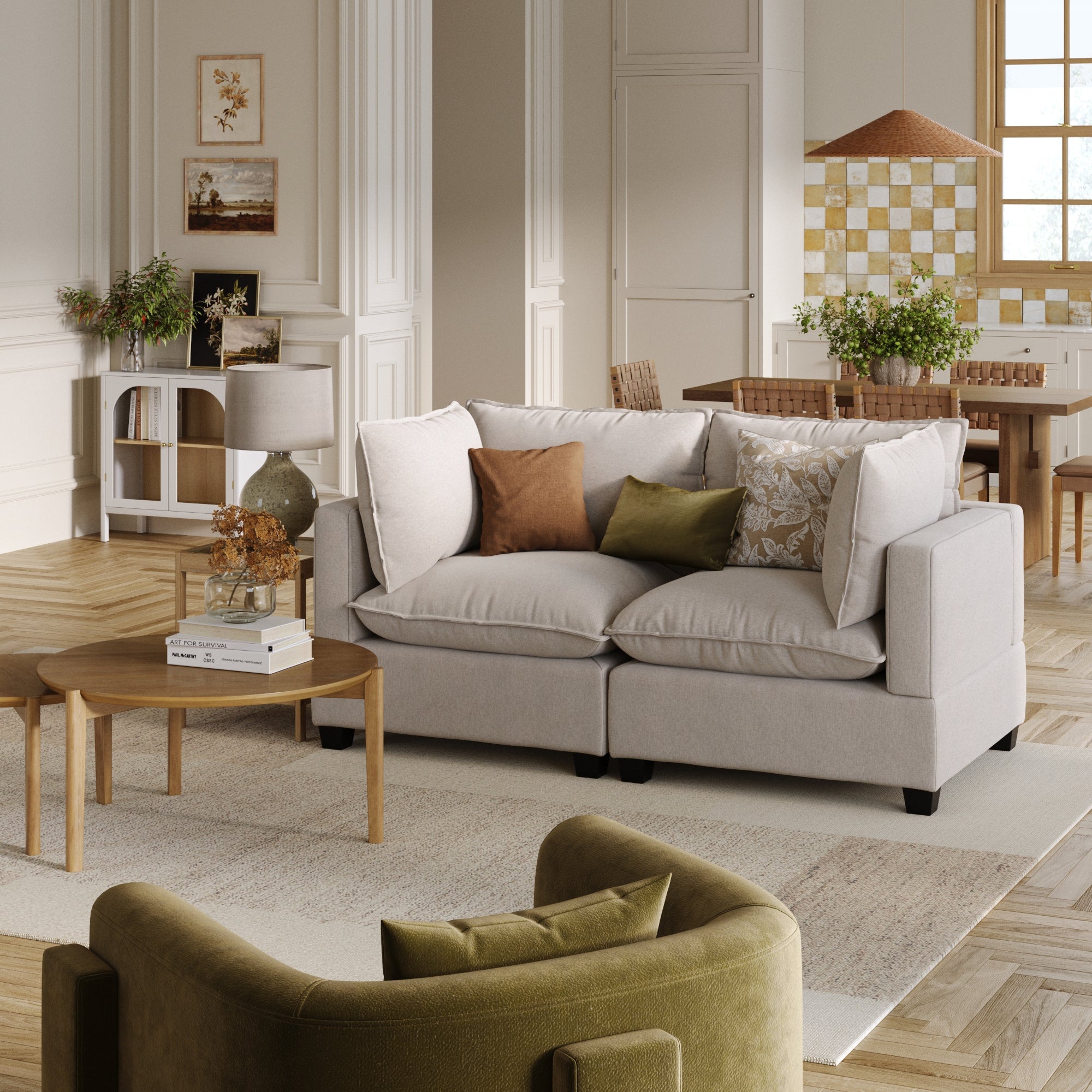 A cozy living room with a Nathan James Soft Fabric Cloud Loveseat Sofa Couch in gray, deep seat cushions, neutral pillows, a wooden coffee table with books and dried flowers, a green armchair, and plant-filled dining area in the background.