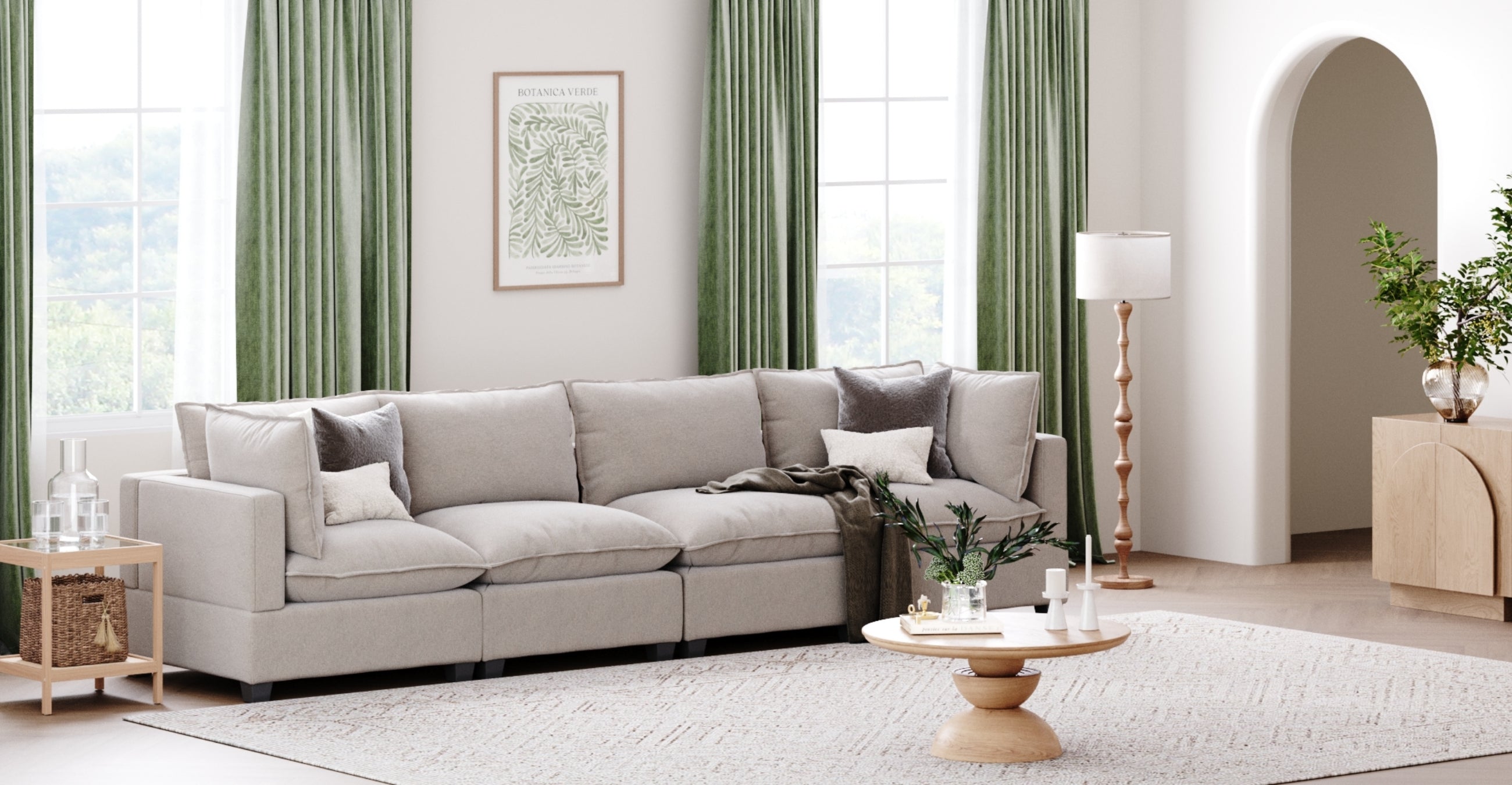 A modern living room with a large light gray sectional sofa, a round wooden coffee table, a floor lamp, green curtains, wall art, and a decorative plant on a sideboard. The room is bright and airy with large windows.