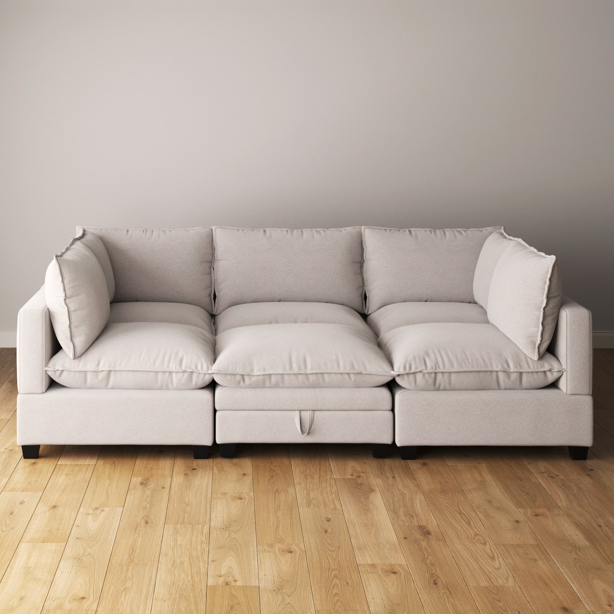 A modern light gray sofa with three seat cushions and plush back pillows sits against a plain beige wall on a light wood floor. The sofa has wide armrests and a soft, inviting appearance.