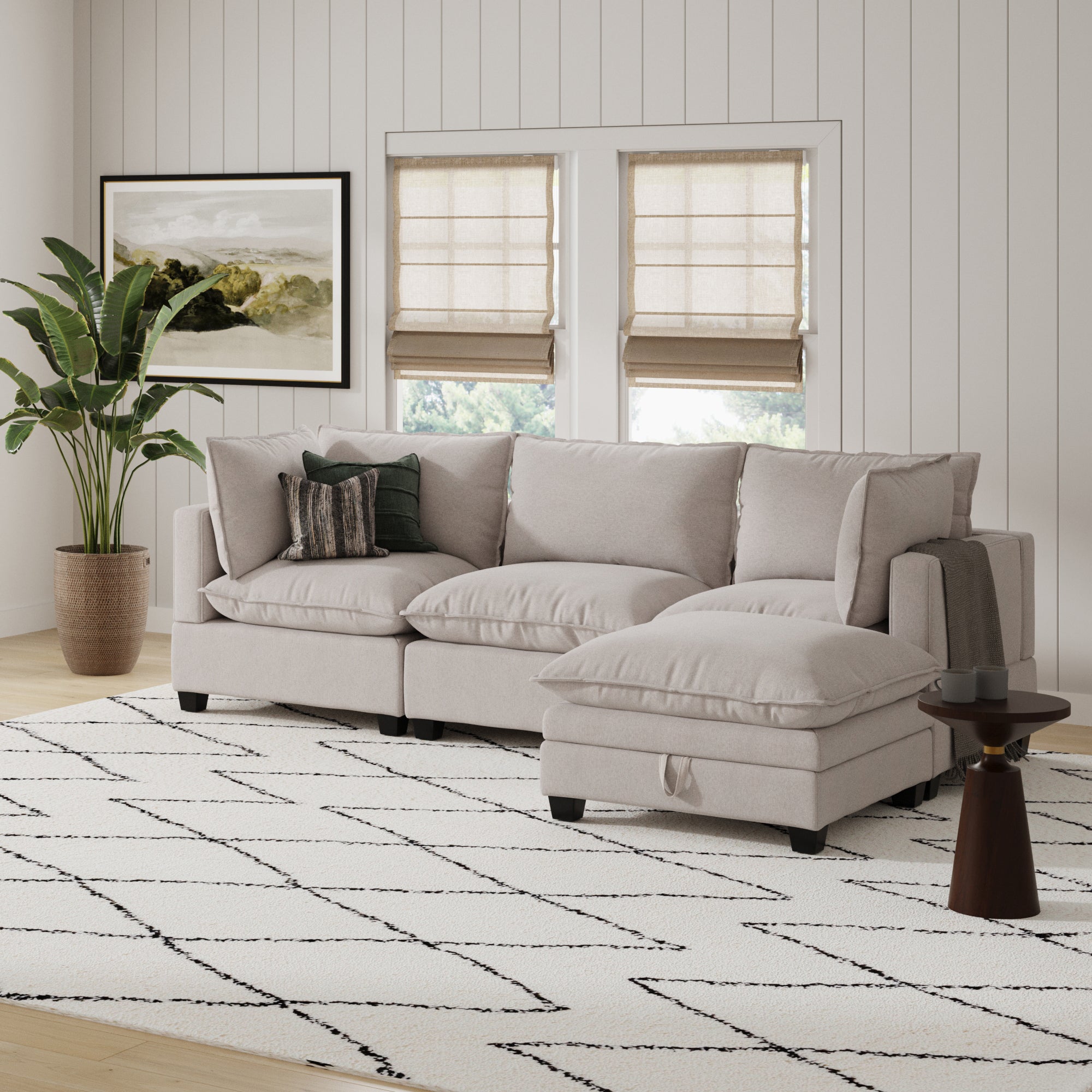 A modern living room has a Nathan James Modern Cloud Sofa Corner Seat in textured gray, a patterned rug, round side table, potted plant, wall art, and two windows with beige roman shades on a white shiplap wall.