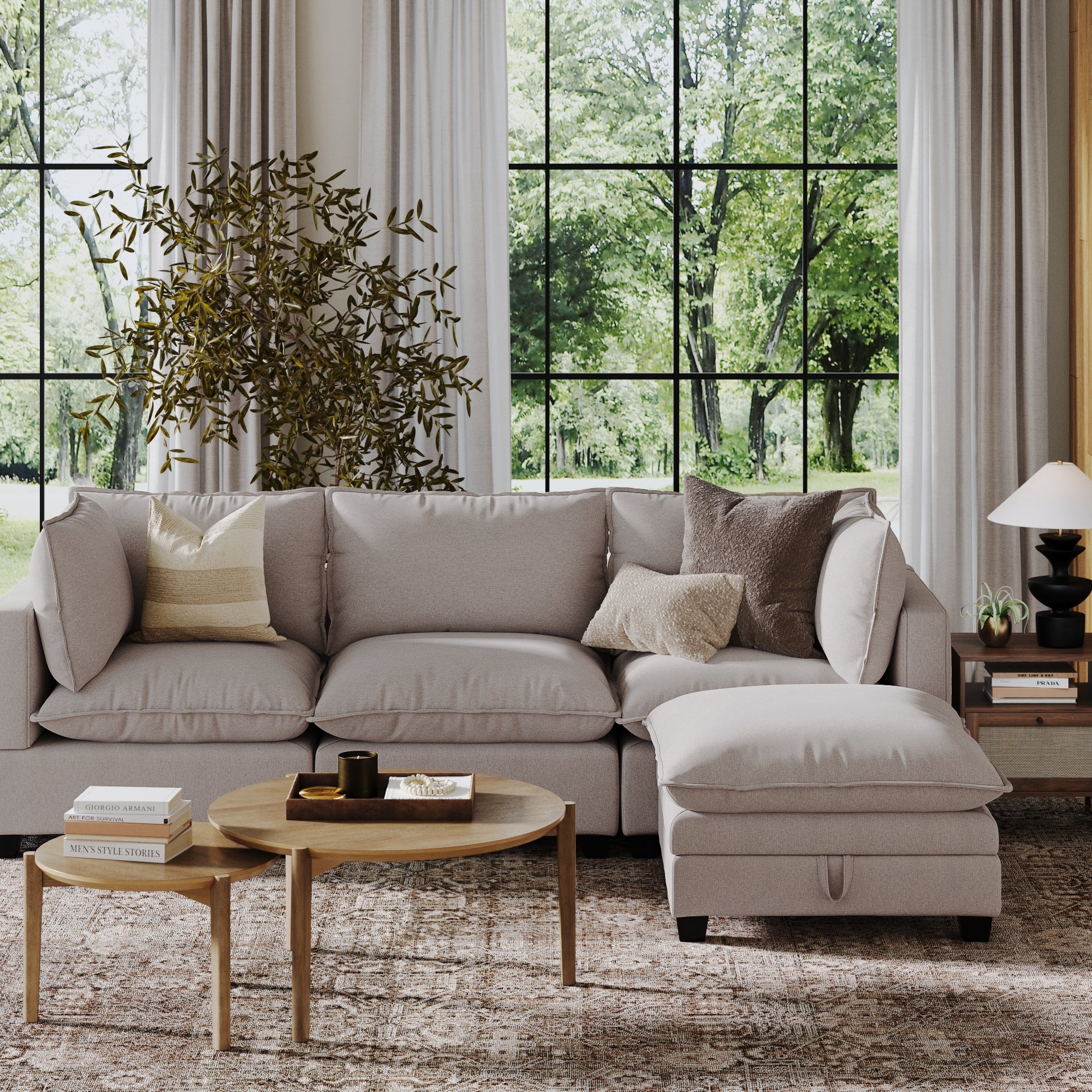 A modern living room features a Nathan James Soft Fabric Hidden Storage Ottoman in gray, decorative pillows, a round wooden coffee table, a potted plant, and large windows with green views. Neutral tones create a cozy, inviting vibe.
