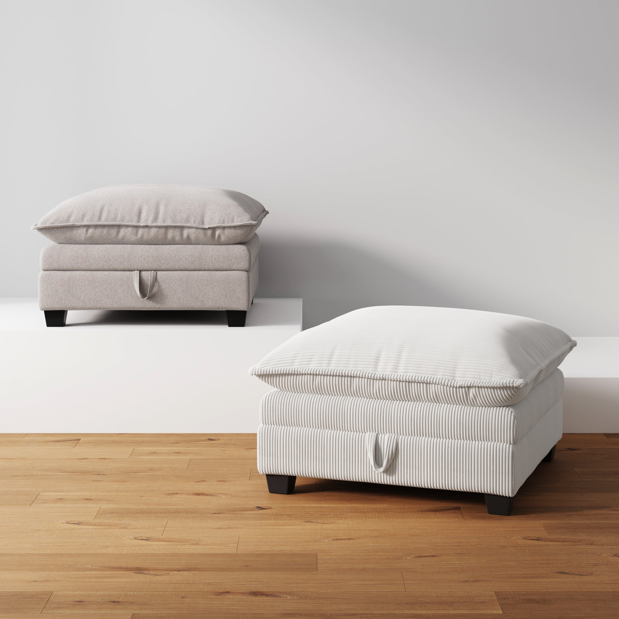 Two cushioned ottomans with pillow-like tops, one beige and one white with subtle stripes, are placed on a wooden floor against a white wall. The ottomans have fabric handles and low black legs.
