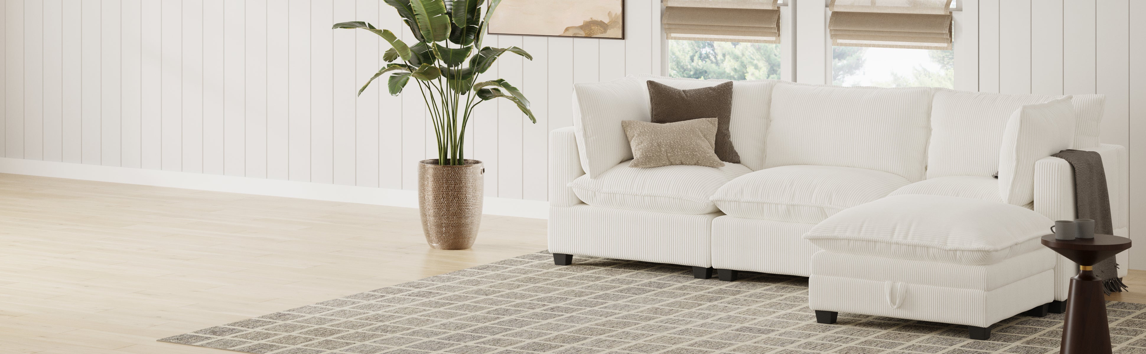 A modern living room features a white sectional sofa with neutral pillows, a potted plant, a small round side table, a patterned rug, and light wood flooring, with natural light coming through two windows.