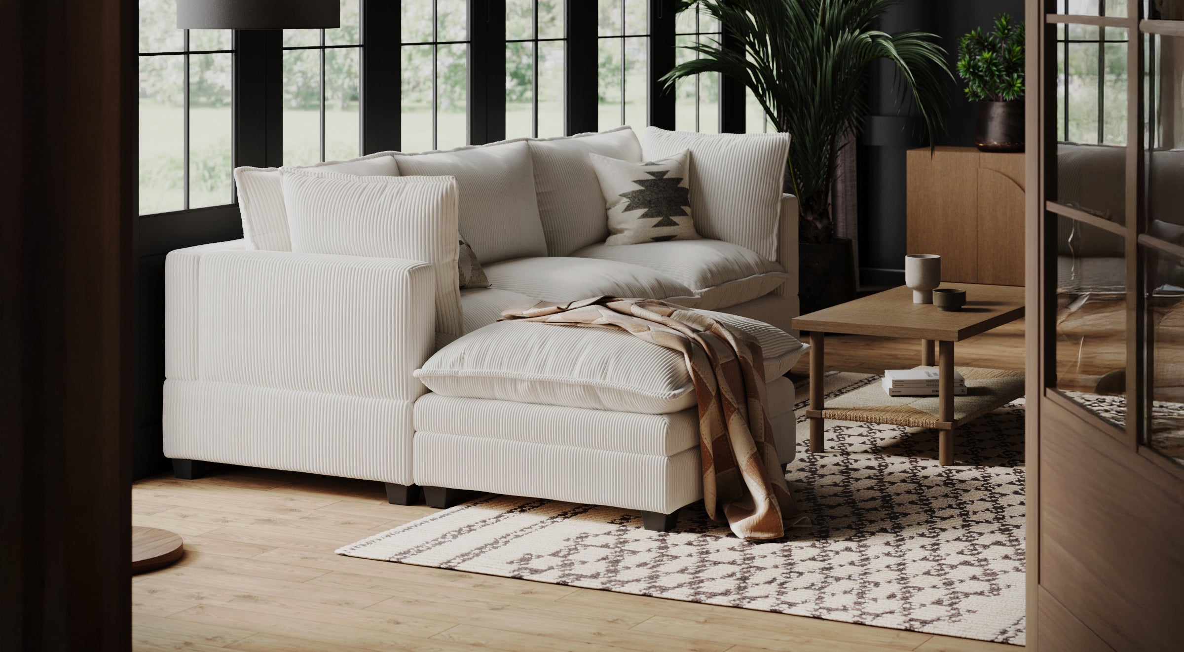 A cozy living room with a white sectional sofa, patterned cushions, a beige blanket, a patterned rug, a wooden coffee table with mugs, a green potted plant, and large windows letting in natural light.