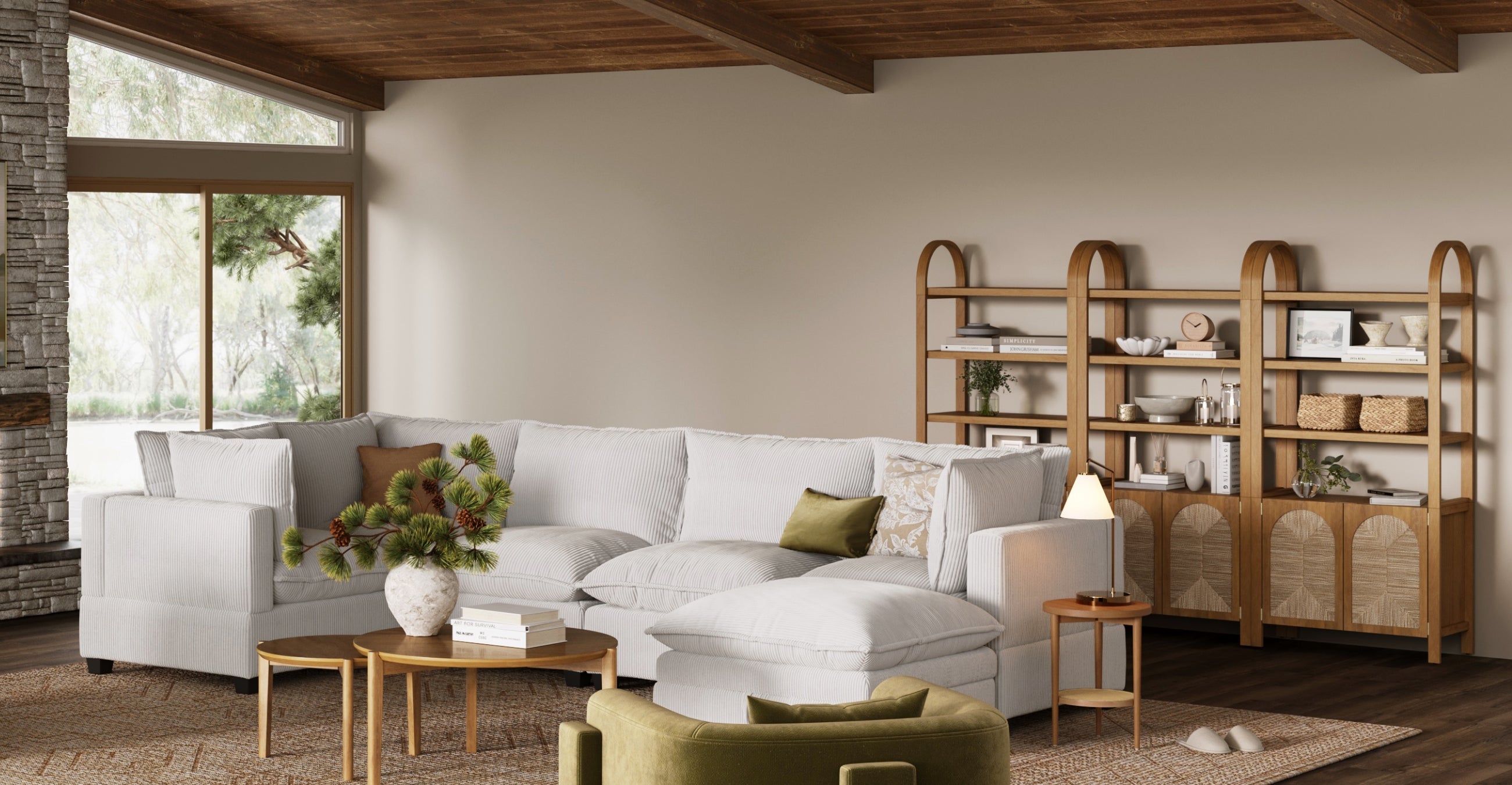 A cozy living room with a white sectional sofa, wooden coffee tables, a green ottoman, a tall shelving unit with books and decor, and large windows letting in natural light.