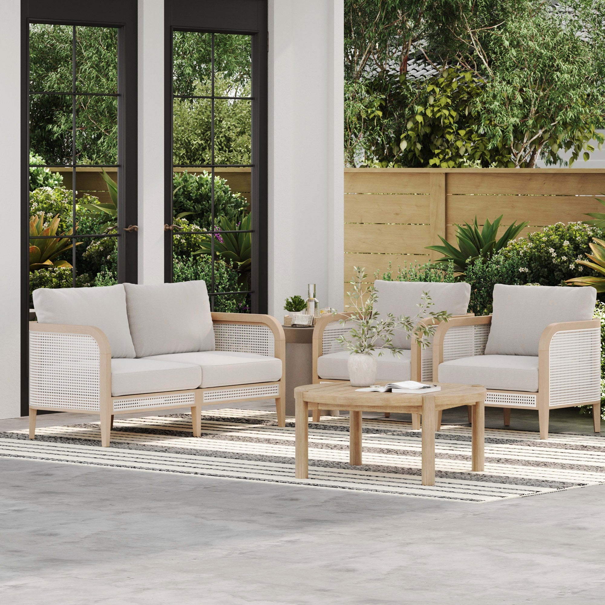 The Nathan James Outdoor 4 Piece Wood Set features a round rattan patio table, two armchairs with beige cushions, and accents of greenery and wood for a stylish, weatherproof outdoor setting.