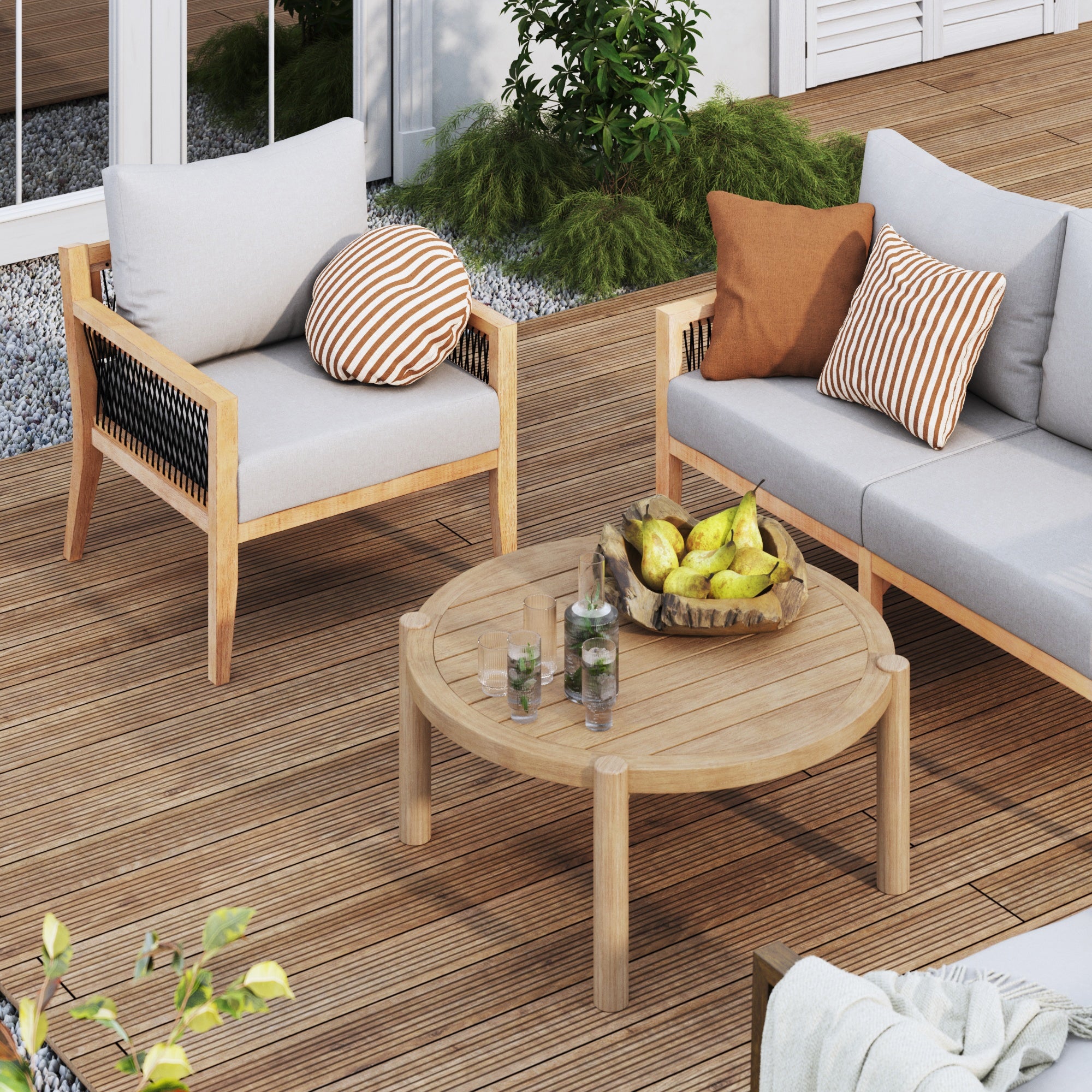 A wooden patio set with cushioned chairs and a round coffee table holding a fruit bowl and two drinks, surrounded by plants on a wooden deck outside a house.