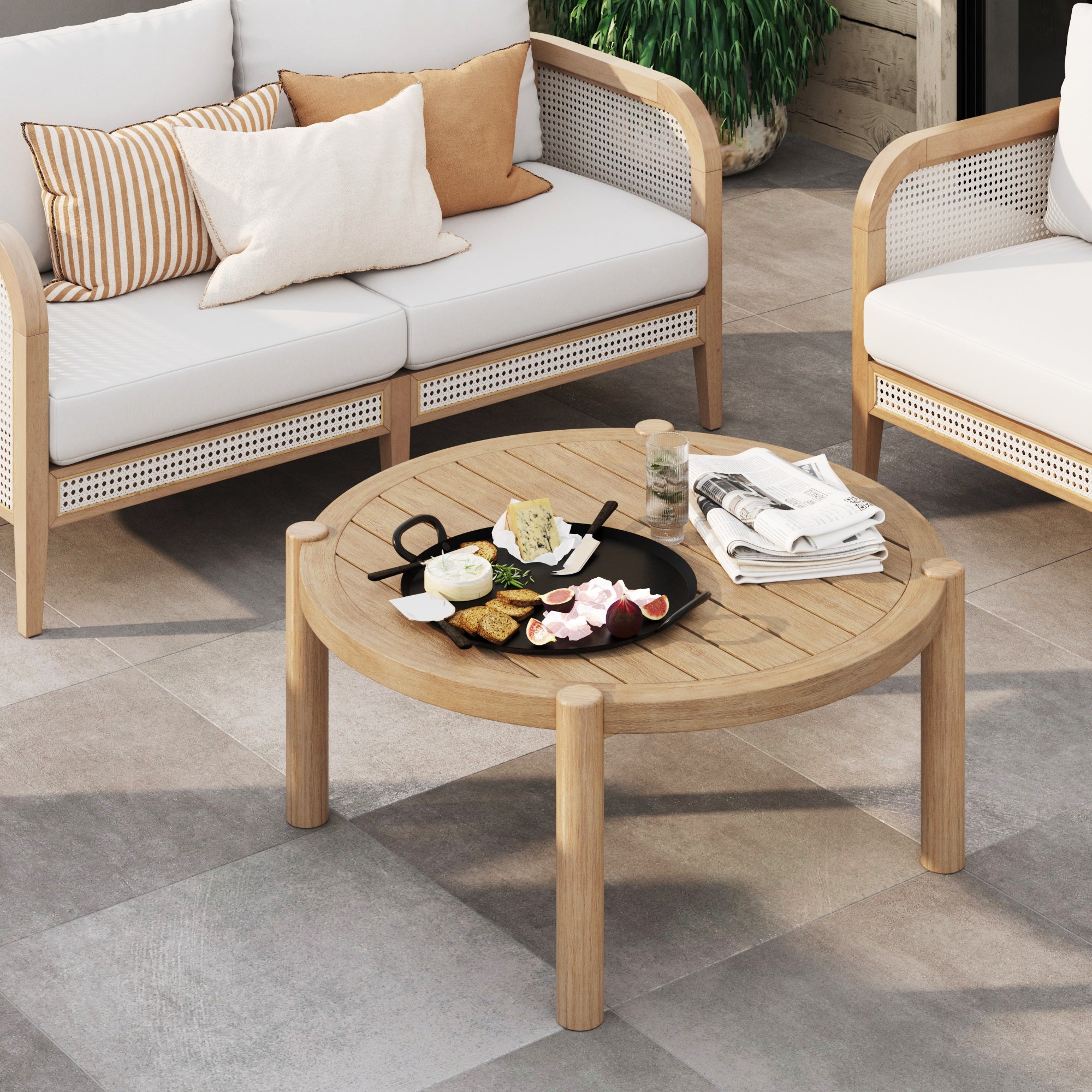 The Nathan James Outdoor Round Wood Coffee Table in weathered brown holds snacks, drinks, and newspapers between two cushioned light sofas with throw pillows on a tiled floor, while a green plant adds freshness in the background.