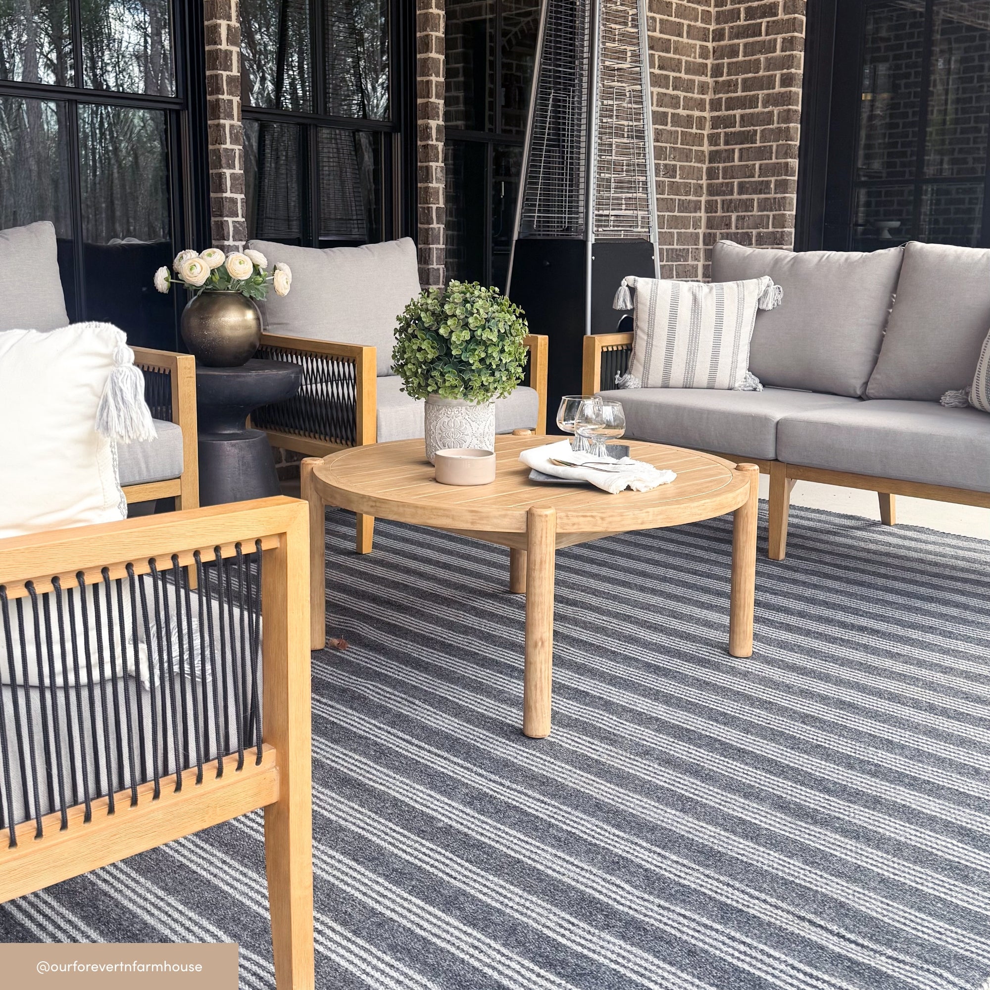 Modern outdoor patio with light wood furniture, gray cushions, a round wooden coffee table with a potted plant, two glasses, and a white napkin on a striped rug. Brick wall and large windows in the background.