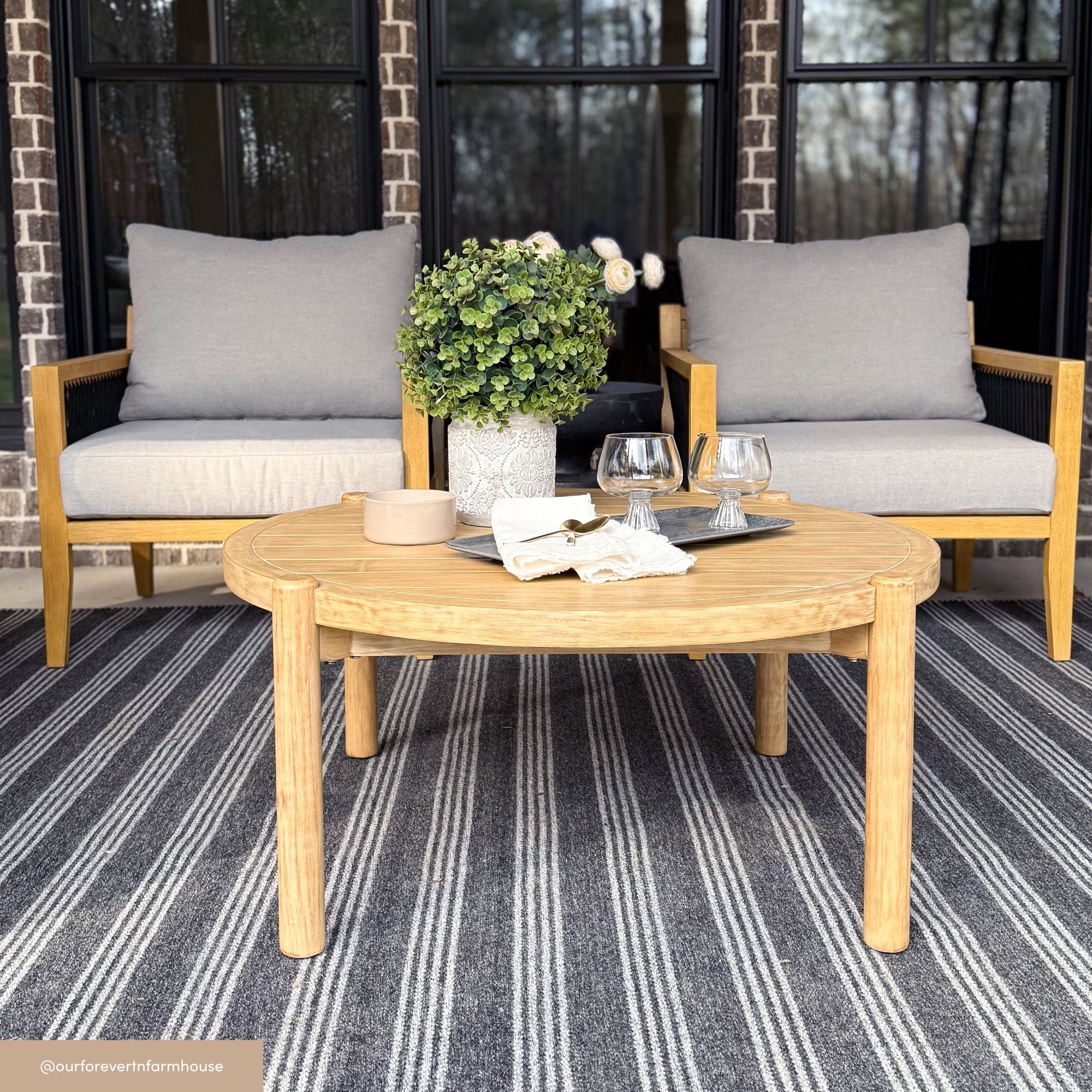 A cozy outdoor seating area with two wooden chairs with gray cushions, a round wooden coffee table holding a potted plant, two glasses, a small dish, and a folded napkin, set on a striped rug in front of large windows.