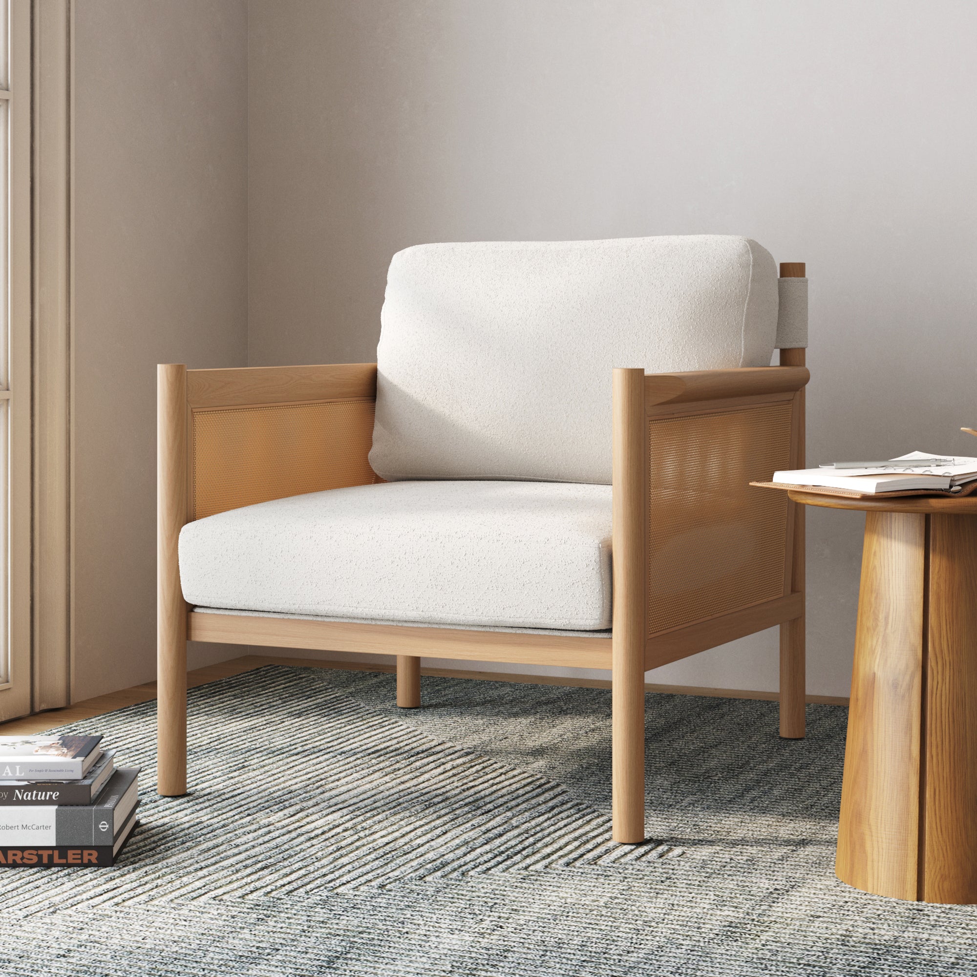The Nathan James Solid Wood Boucle Accent Chair with rattan arms and raw oak finish sits on a patterned rug beside a round wooden side table, books stacked nearby in a softly lit room.