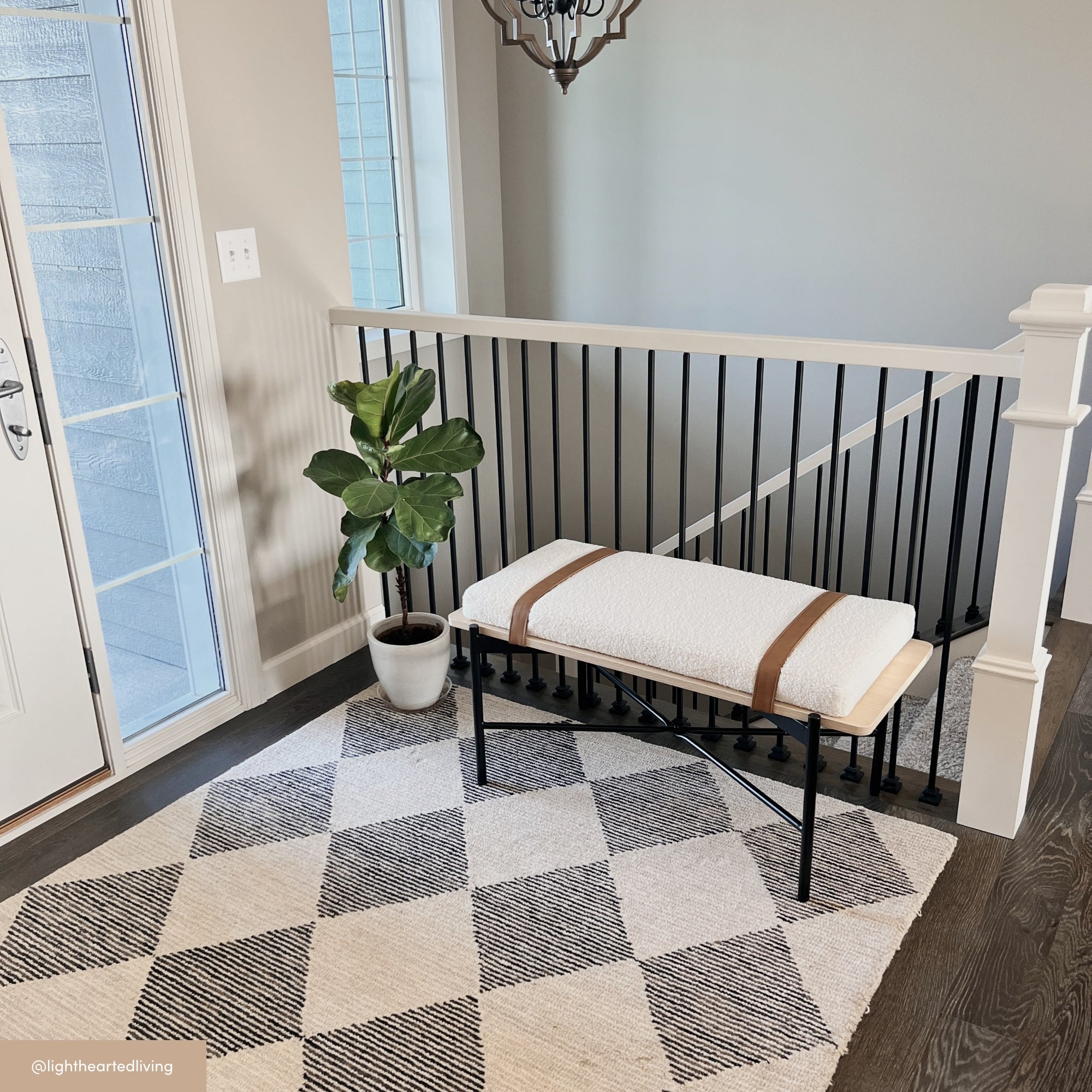 A modern entryway with a black-and-white checkered rug, a light-colored bench with a cushion and leather straps, a potted plant, and a staircase railing with black balusters. Natural light streams through nearby windows.