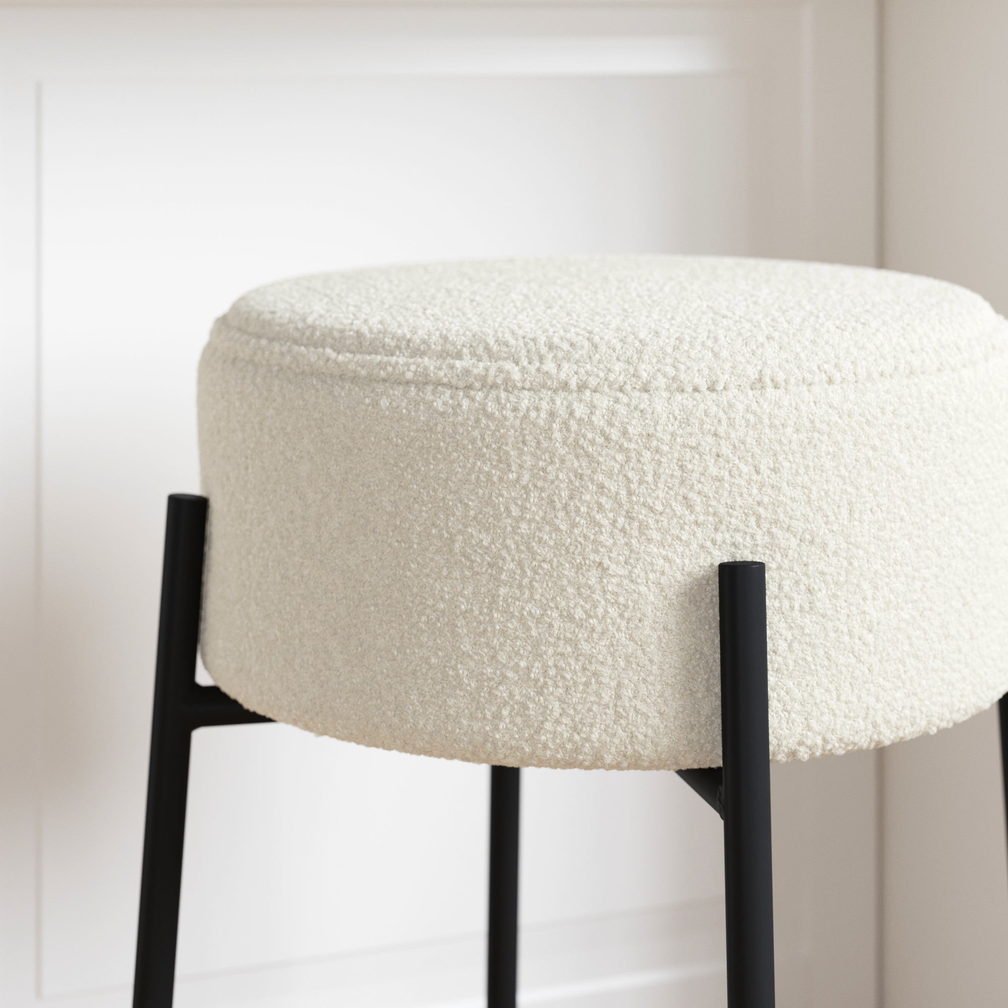 A close-up of a round, cream-colored, textured stool with a soft boucle fabric seat and black metal legs, placed in front of a white paneled wall.