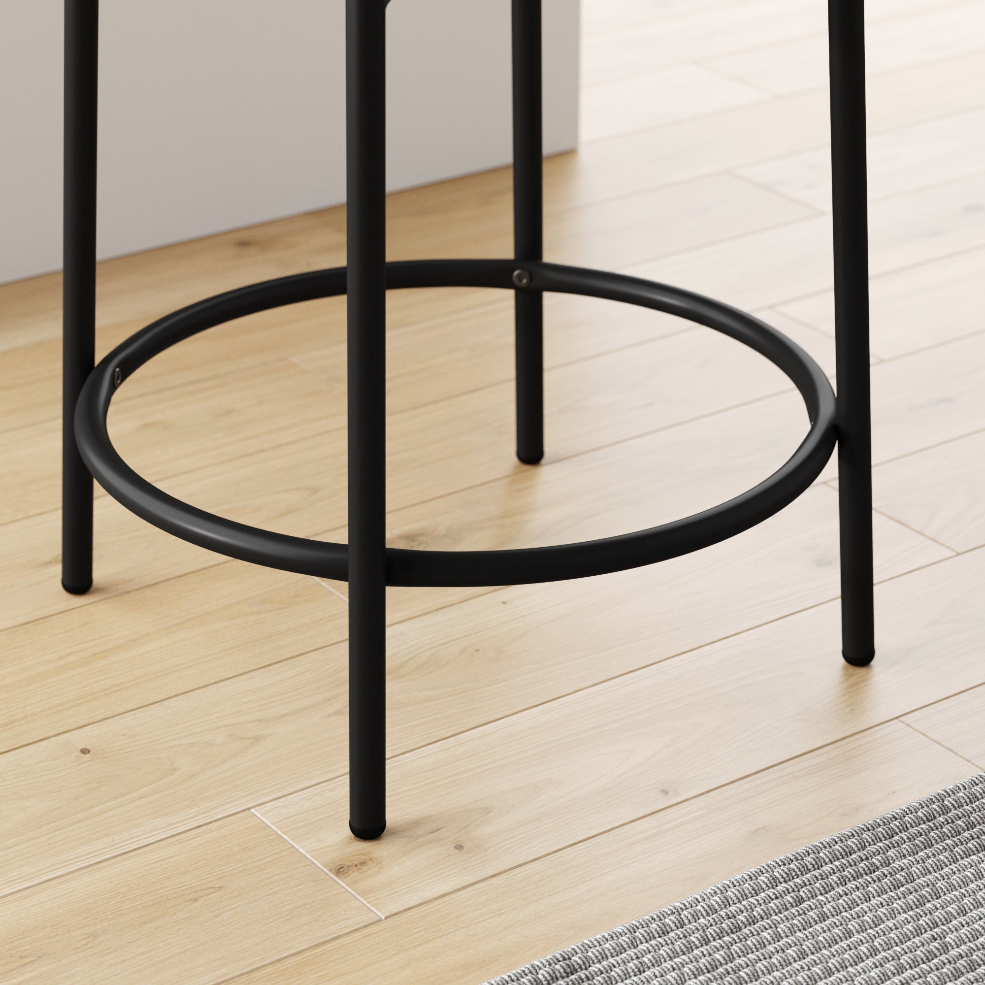 Close-up of the black metal legs and circular footrest of a bar stool set on a light wood floor, next to a gray textured rug and white furniture in a modern interior.