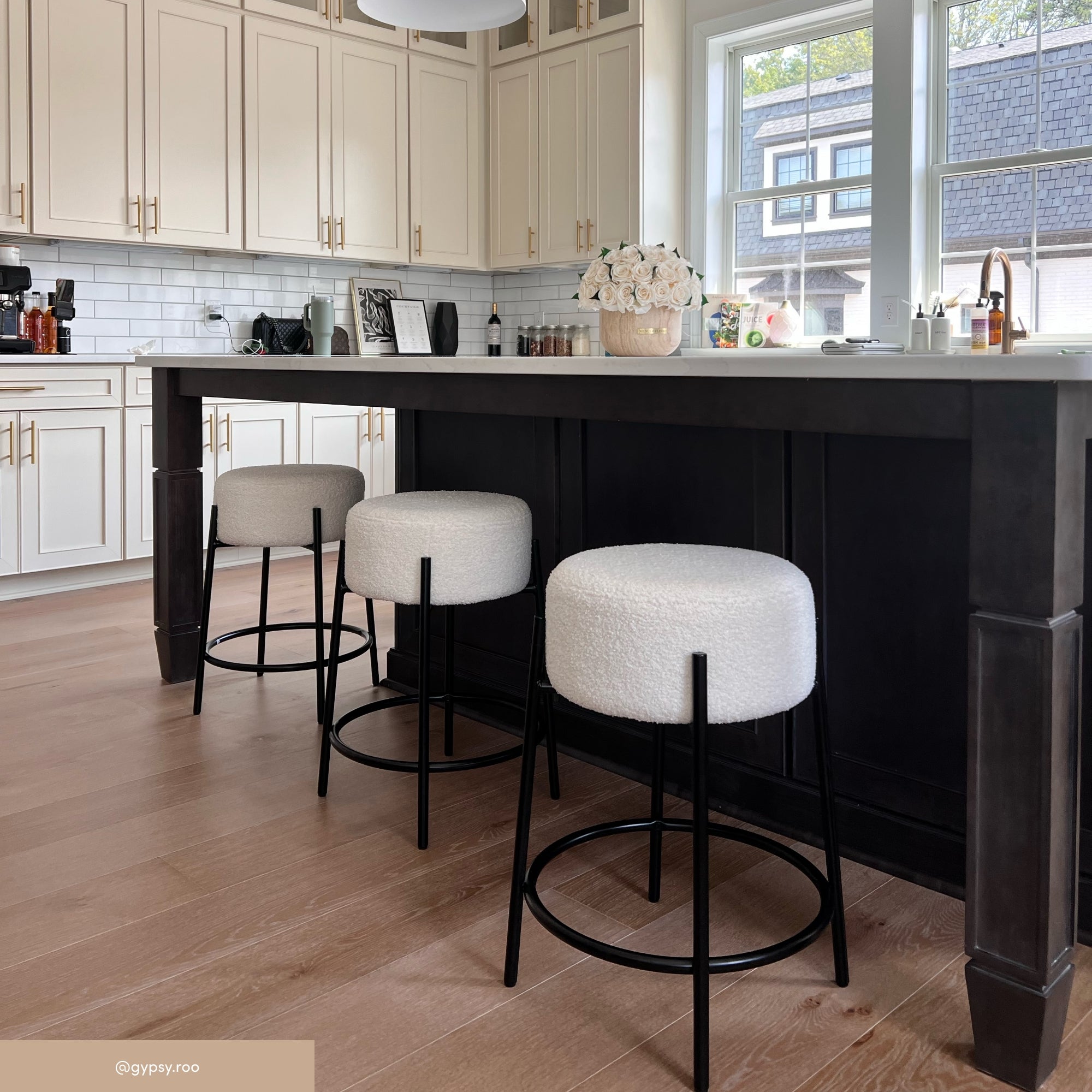 A modern kitchen features a large dark island with three white cushioned stools, light wood floors, cream cabinets, subway tile backsplash, and a vase of flowers on the counter near a bright window.