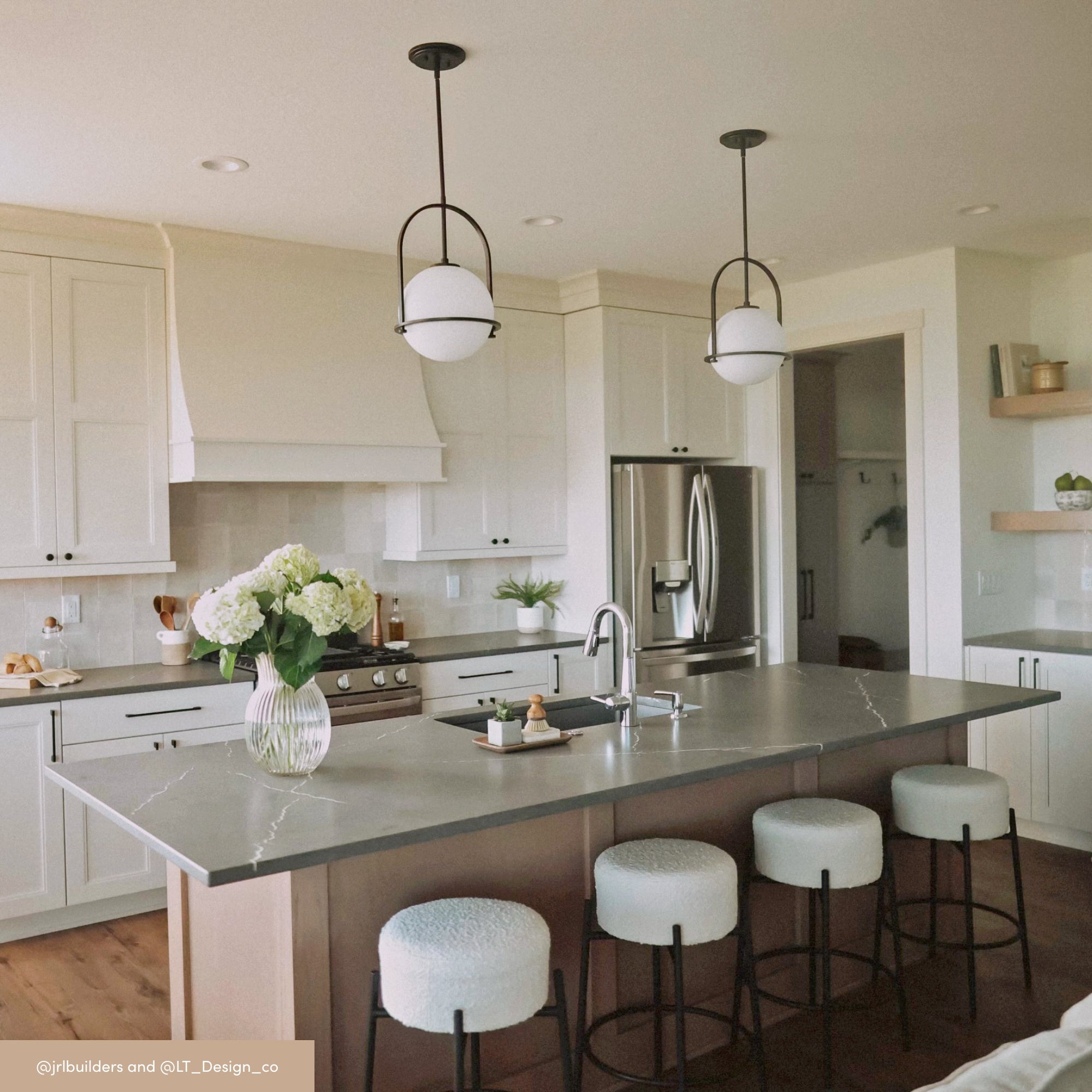 A modern kitchen with white cabinets, stainless steel appliances, and a large gray island. Four cushioned stools are at the island, and two pendant lights hang above. A vase of flowers sits on the counter.