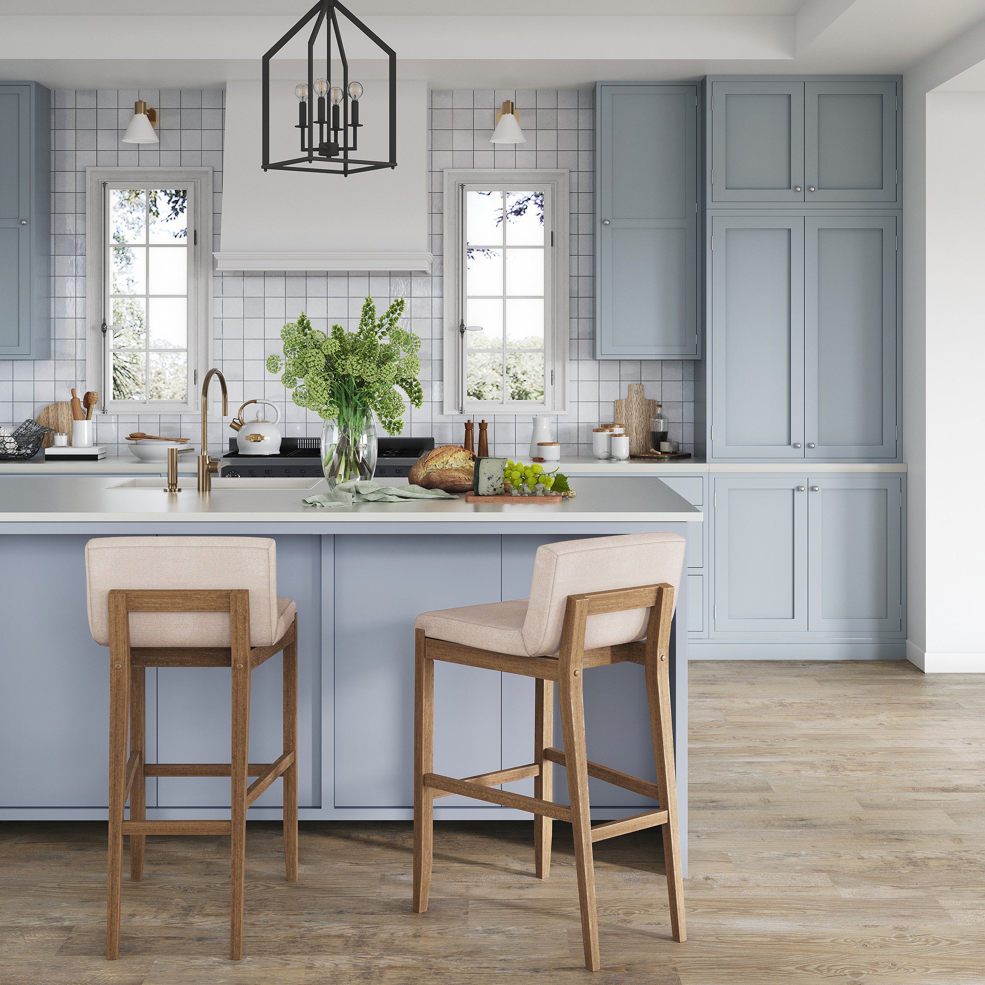 A modern kitchen with light blue cabinets, a white island, two beige bar stools with wooden legs, a vase of green flowers, and large windows letting in natural light.
