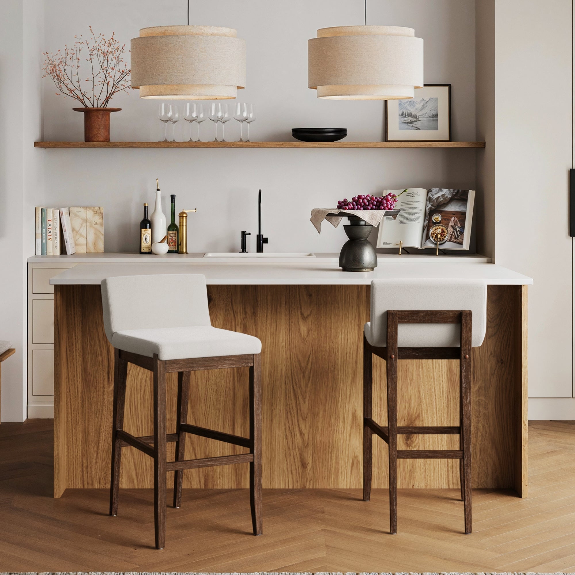 Modern kitchen with a wooden island, two Nathan James Exposed Wood Back Bar Stools (Dark Brown), pendant lights, a shelf with glasses, bottles, decor, vase, and a bowl of fruit on the countertop.