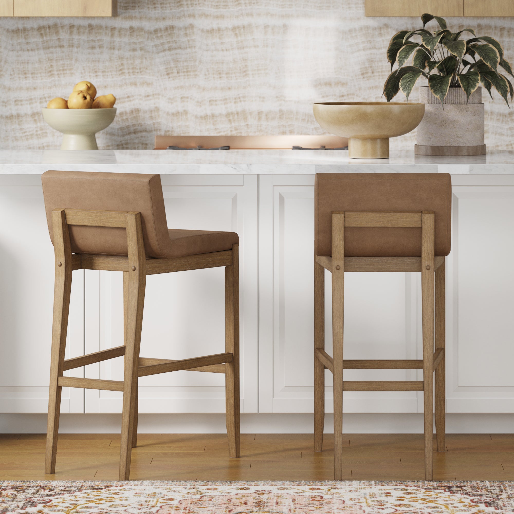 Two Nathan James Exposed Wood Back Bar Stools in light brown are at a white kitchen counter with faux leather cushions. On the counter are a fruit bowl, decorative bowl, and potted plant. Light cabinets and a patterned wall complete the look.