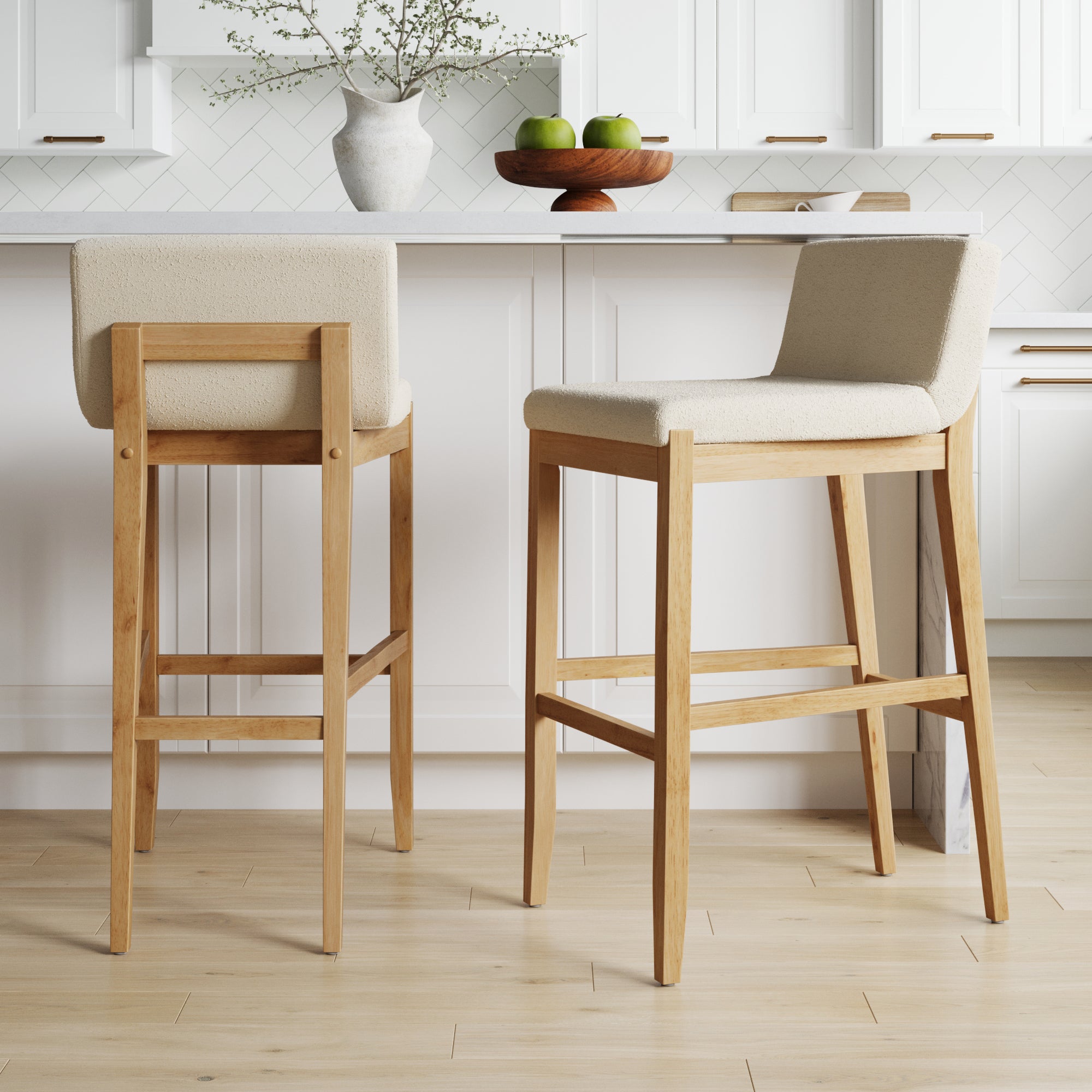 Two light wood bar stools with beige upholstered seats and low backs are placed in front of a white kitchen counter. On the counter, there is a vase with branches and a fruit bowl with green apples.