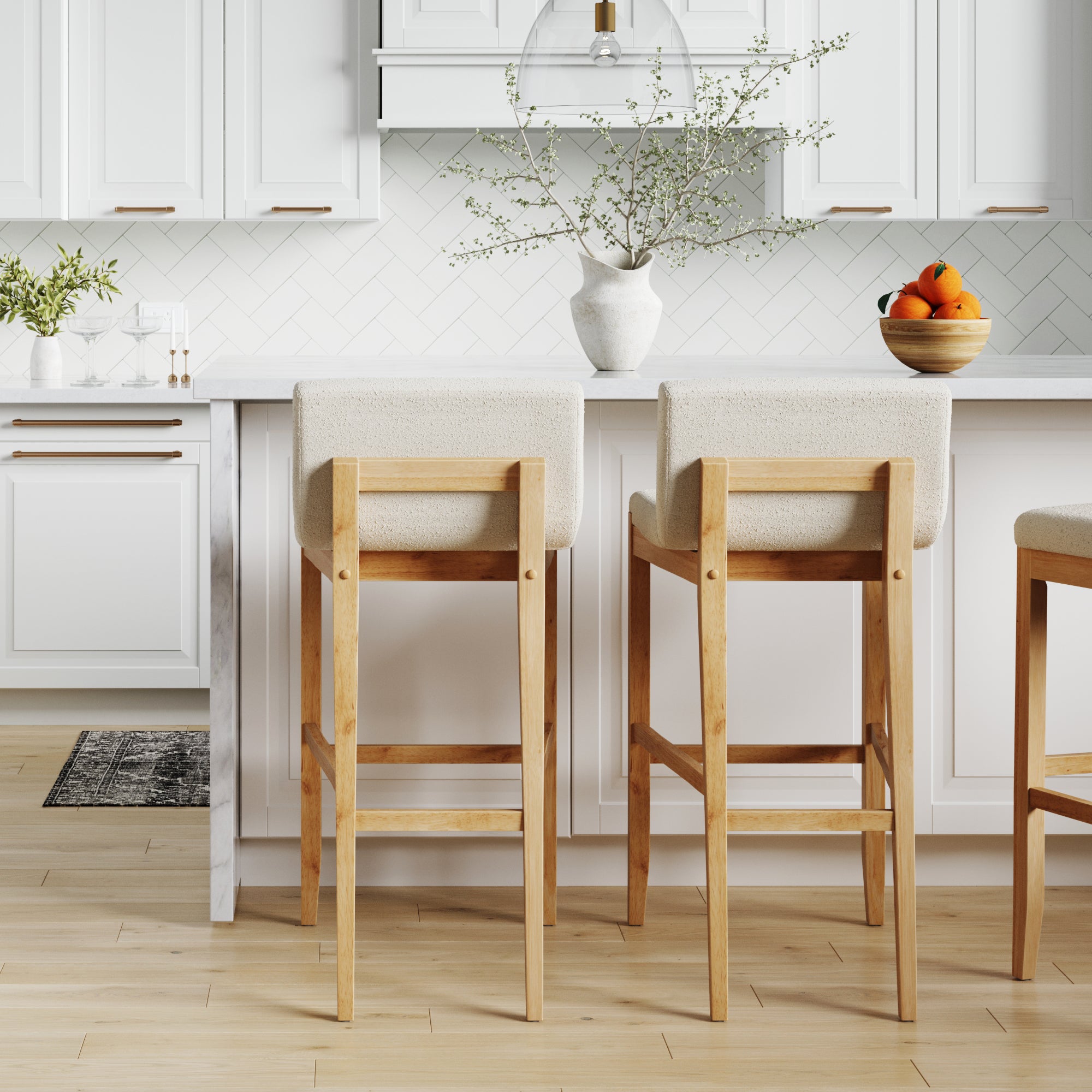 A modern kitchen with white cabinets, a white countertop, and light wood flooring. Two wooden bar stools with white seats face the counter. A vase with branches and a bowl of oranges sit on the counter.
