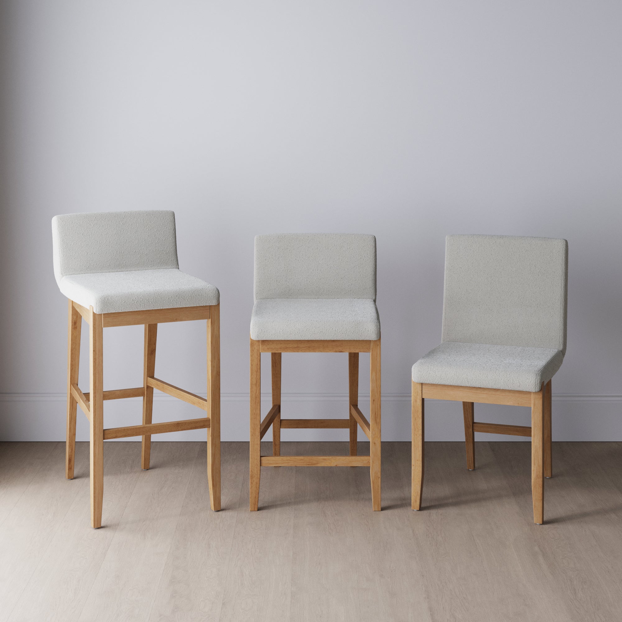 Three wooden chairs with light gray upholstered seats and backs are arranged in a row, each at a different height, against a plain light gray wall and light wood floor.